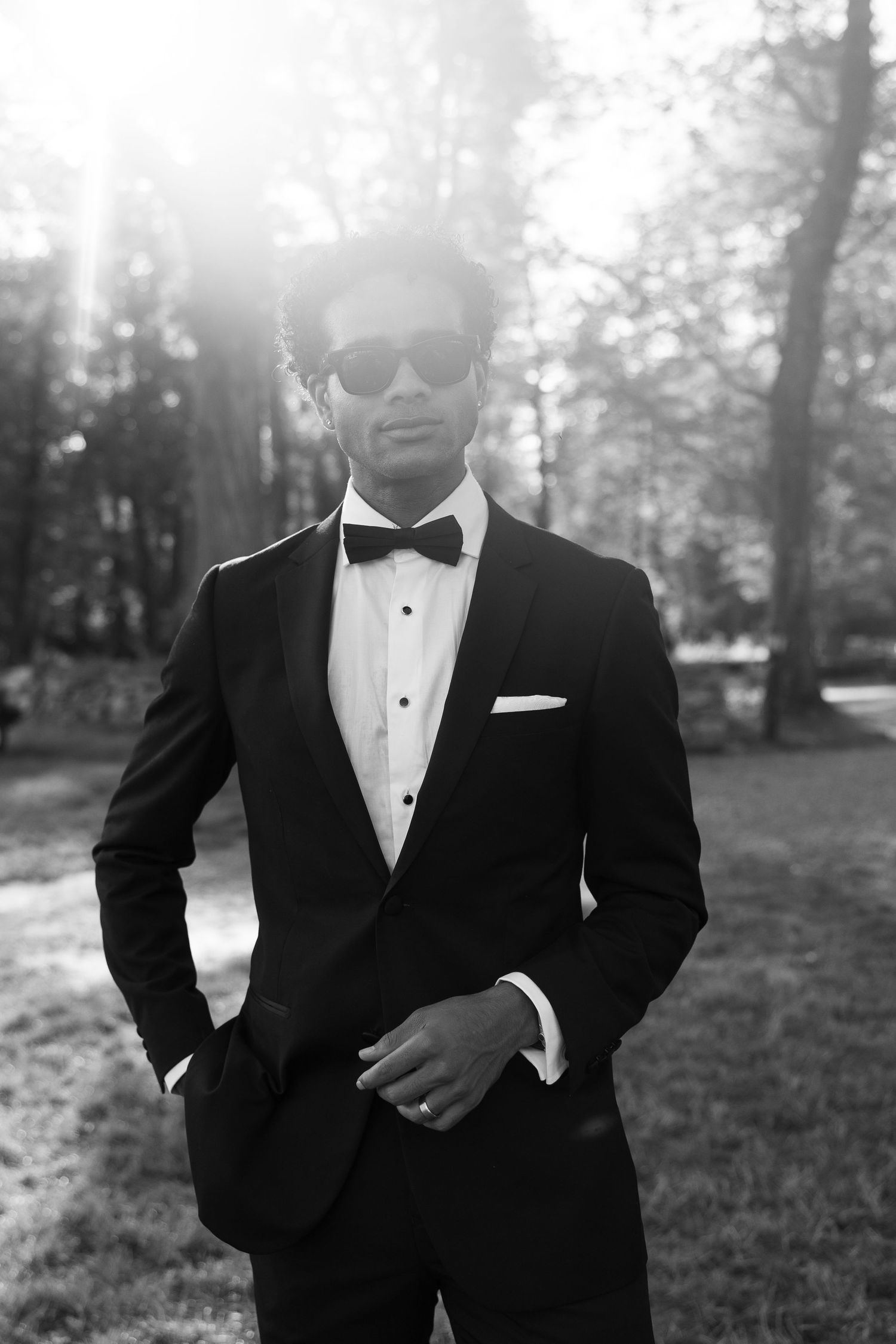 Elegant man in black tuxedo and sunglasses posing outdoors in a sunlit park.