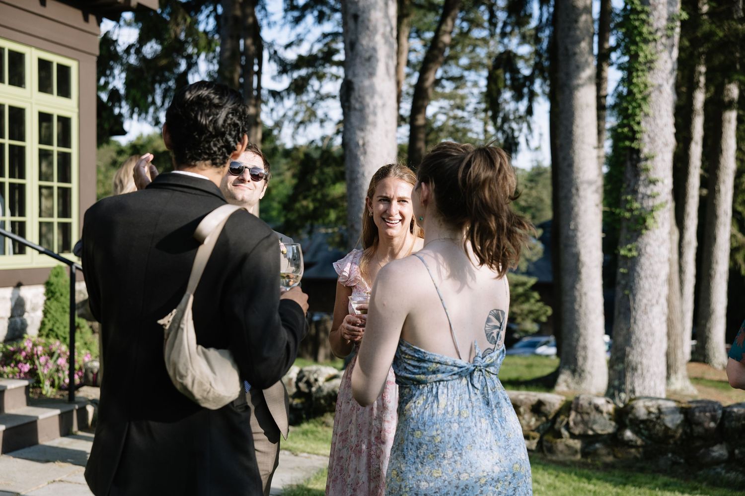 Guests in elegant attire socializing outdoors at a summer garden party, holding drinks among tall trees.
