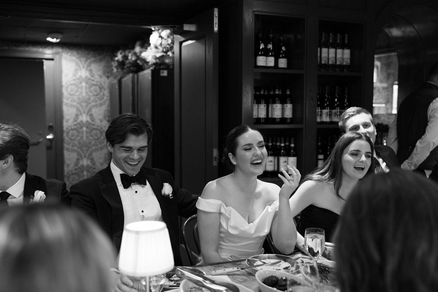 Black and white wedding reception photo of bride and groom laughing with guests at elegantly set dinner table.