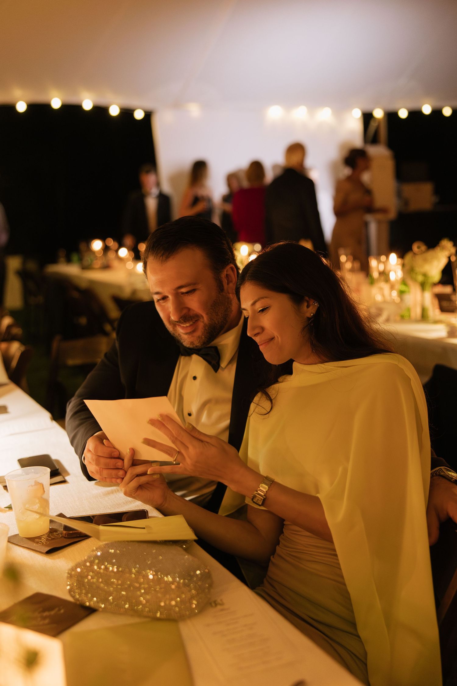 Couple reads a card together at a candlelit wedding reception under a tent with string lights.