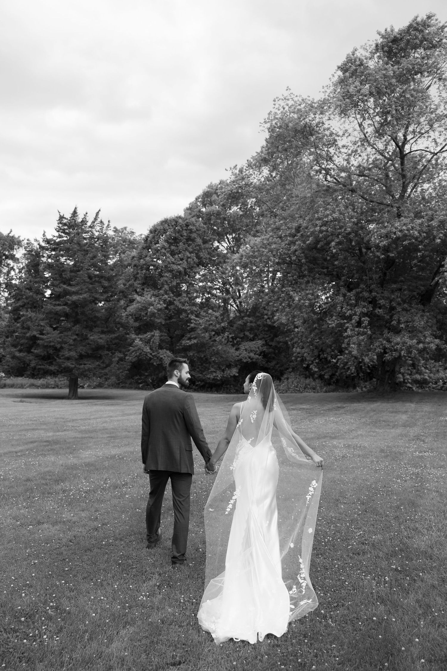 Black and white photo of a bride and groom holding hands walking through a lush green park, brides veil flowing behind her.