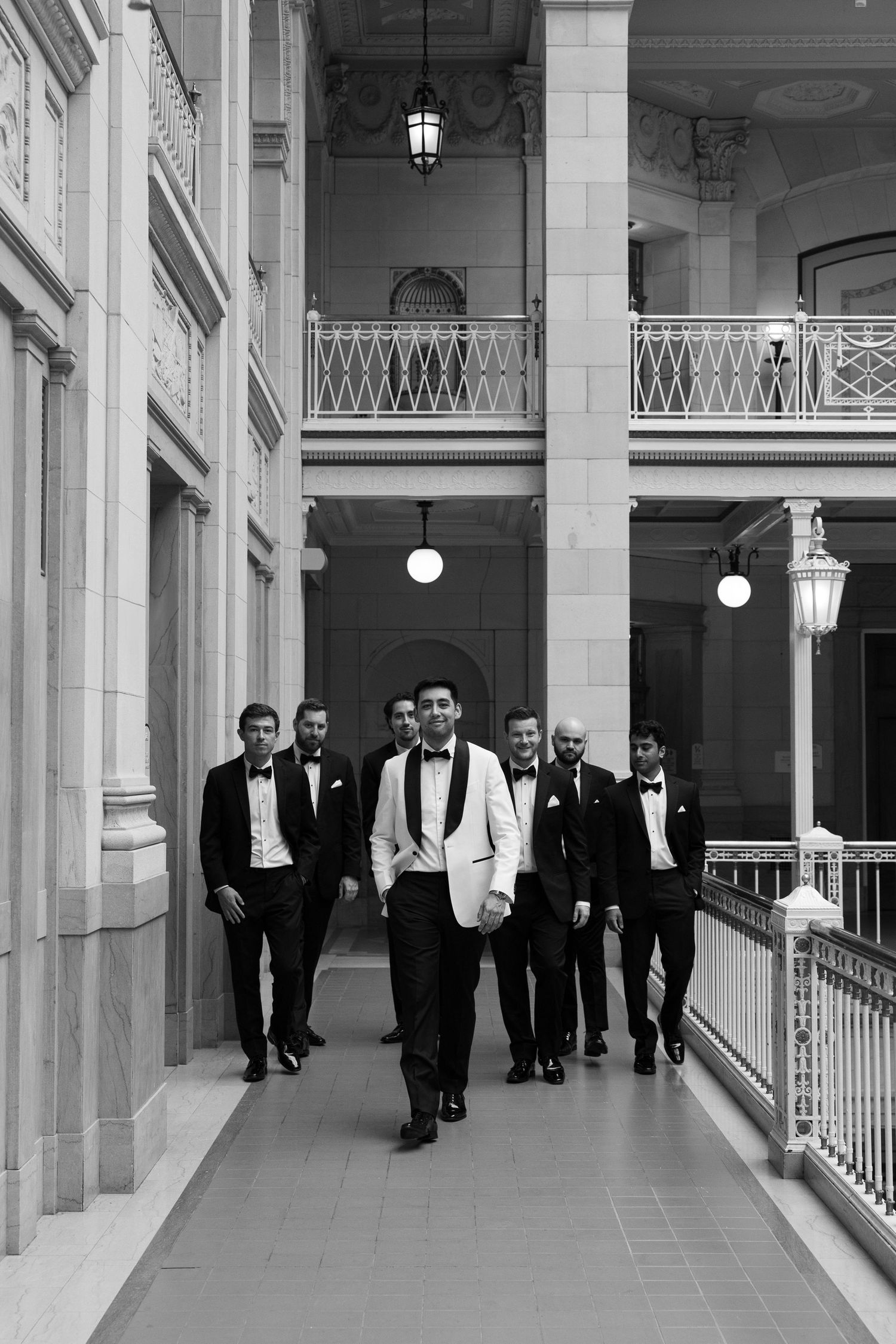 Groom leads groomsmen in tuxedos through a grand classical building corridor in black and white photo.