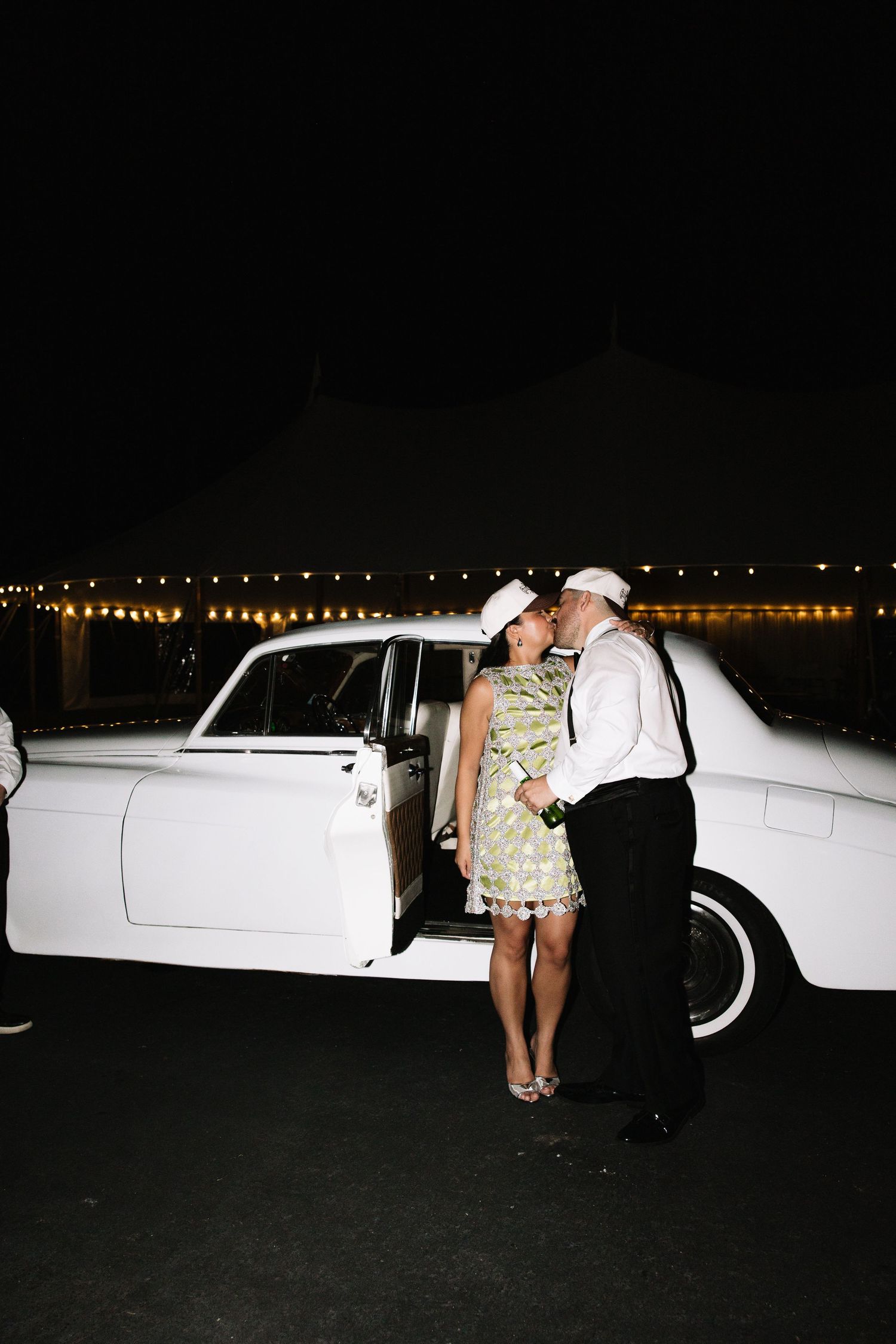 Newlyweds kiss beside vintage white classic car at night with string lights glowing at wedding reception.