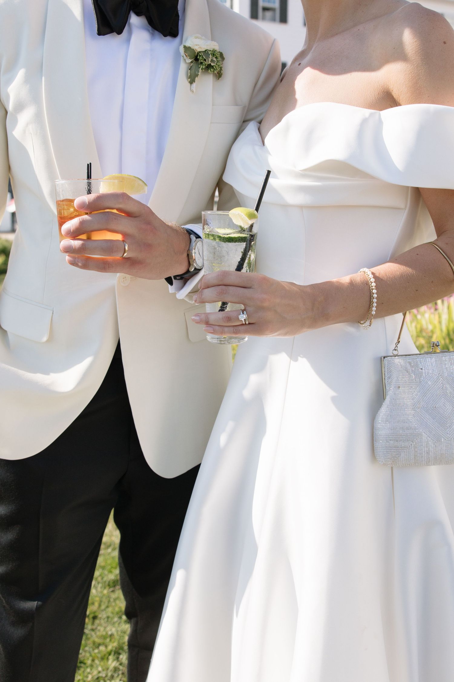 Bride and groom holding cocktails at outdoor wedding, bride in off-shoulder white gown with diamond ring, groom in white tuxedo.