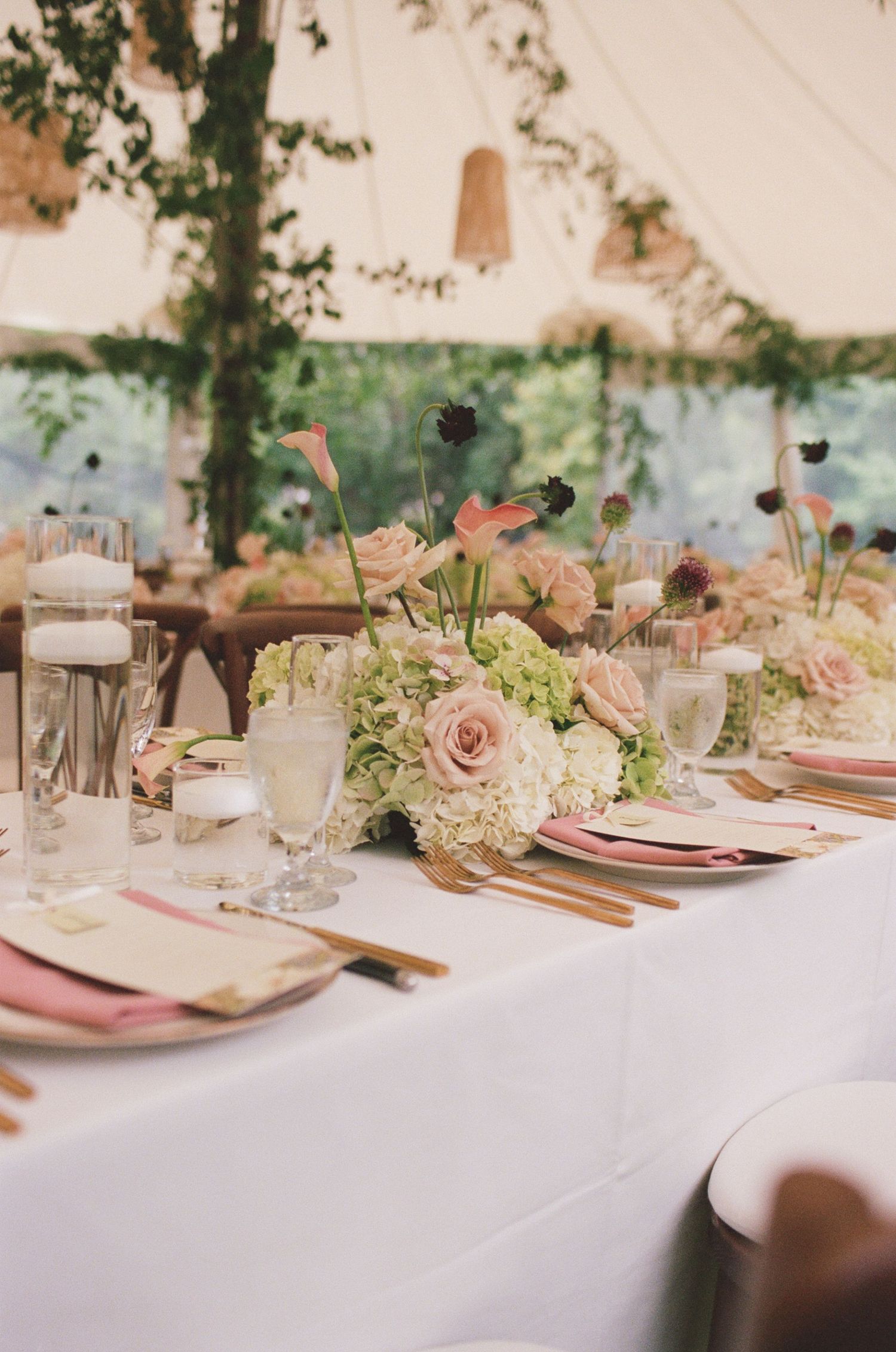 Elegant wedding reception table with blush floral centerpieces, pink napkins, gold cutlery, and candles under a white tent.