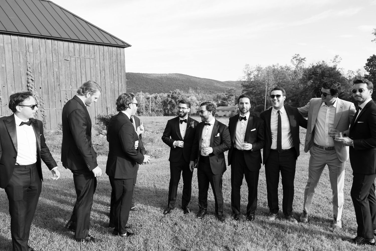 Black and white photo of groomsmen in tuxedos laughing outdoors near a barn on a wedding day.