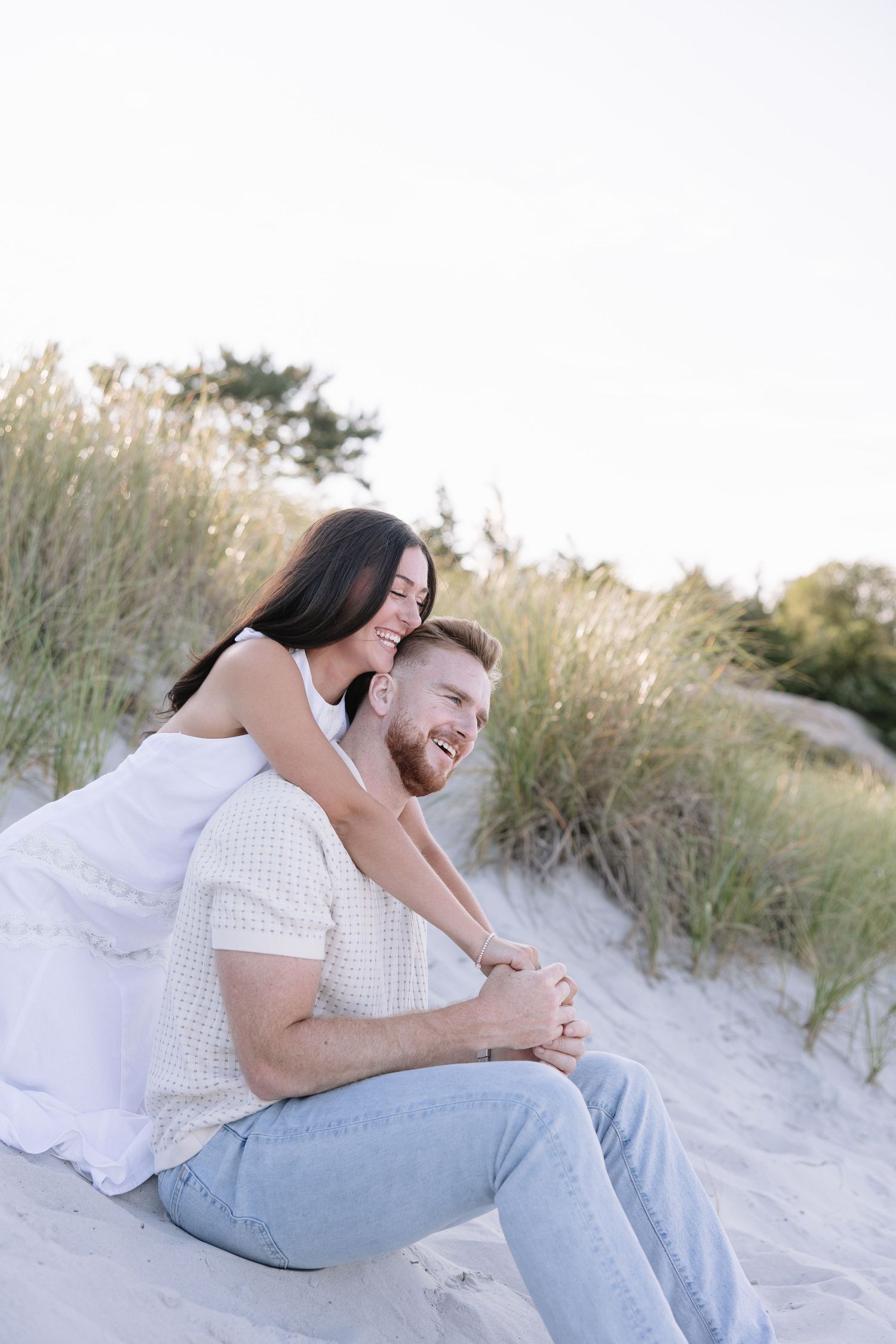 Happy couple sitting on sandy beach dunes, woman in white dress embracing smiling bearded man in casual summer outfit.