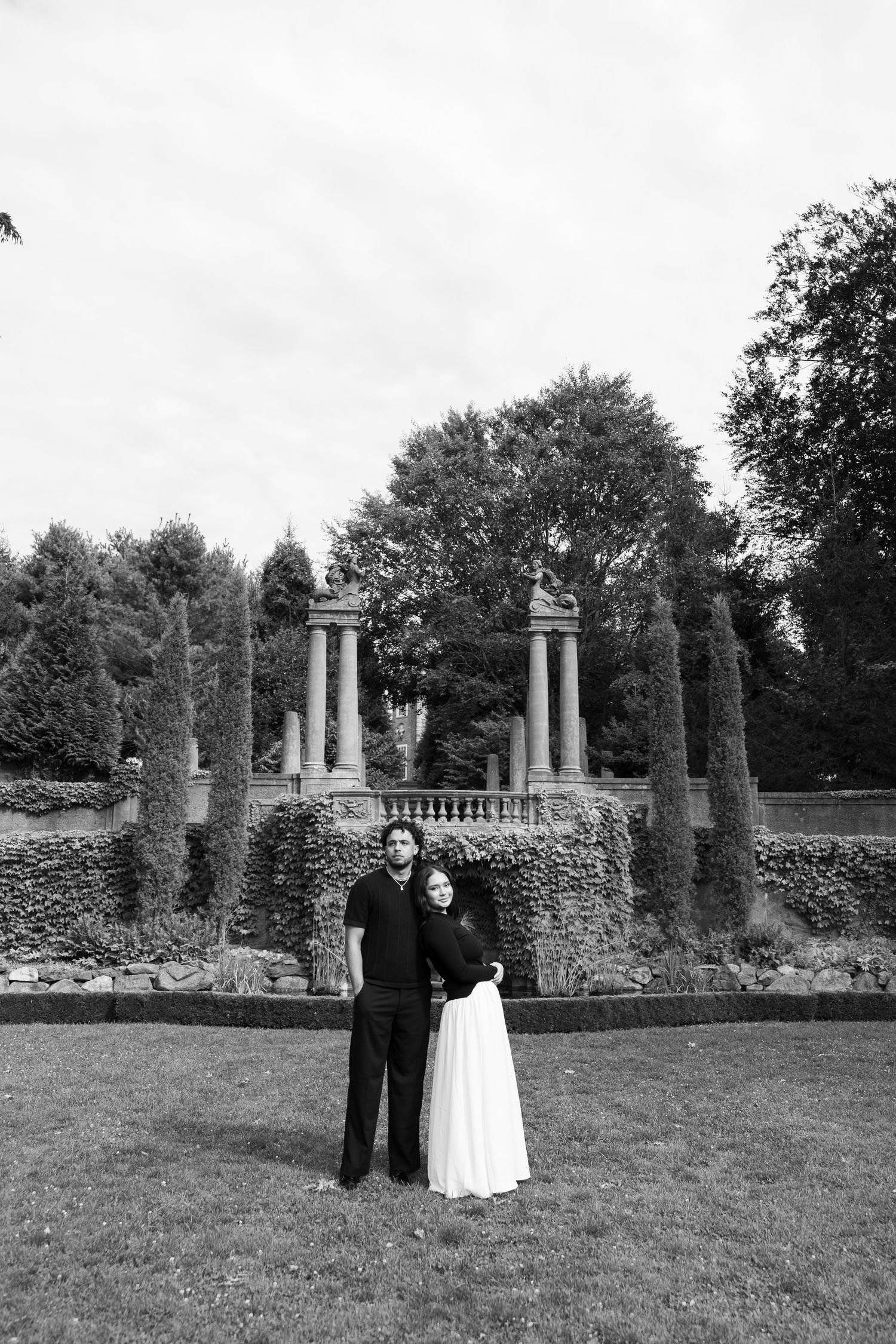 A black and white photo of a couple standing before classical stone columns in a garden.