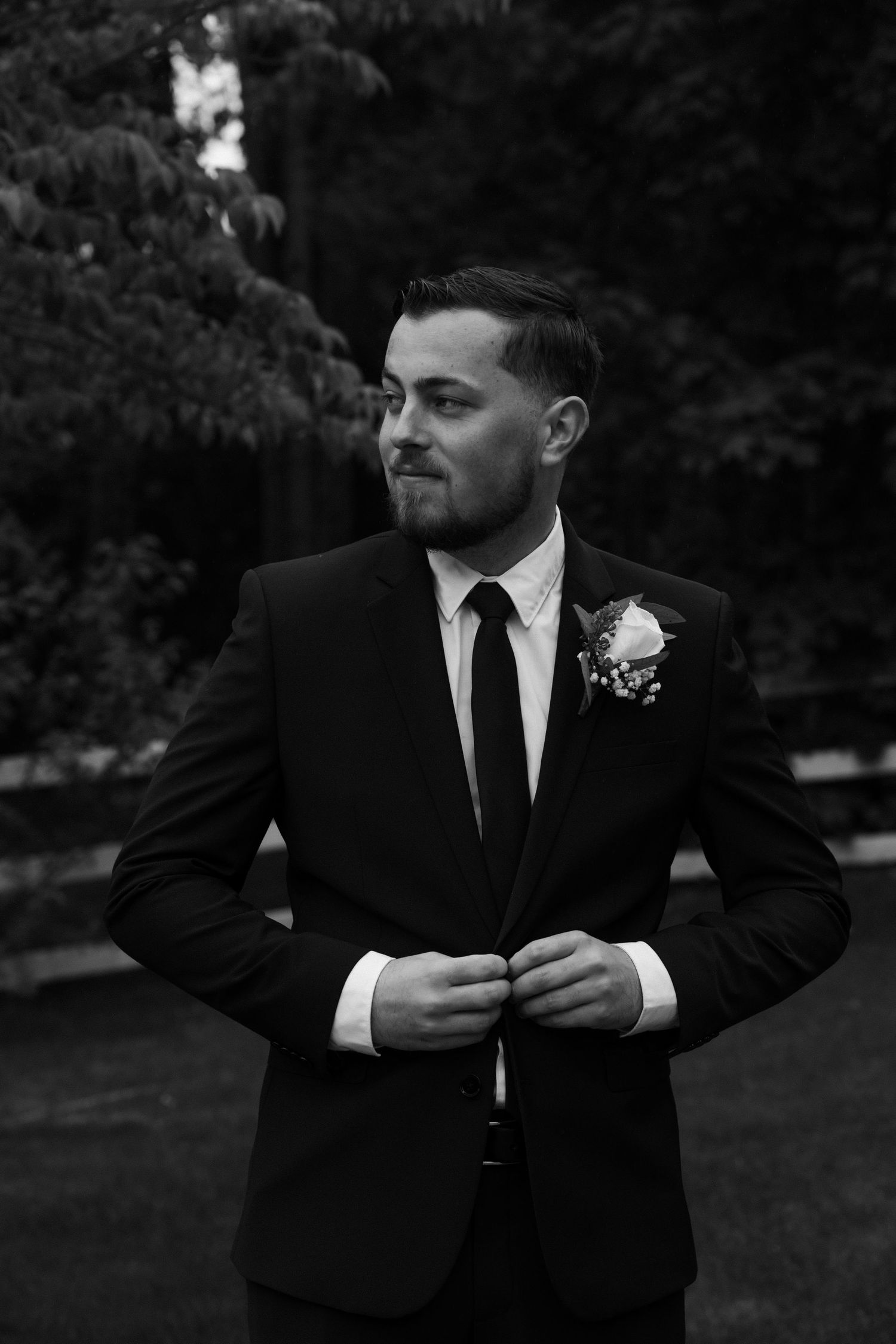 Man in black suit and tie adjusts his jacket outdoors.