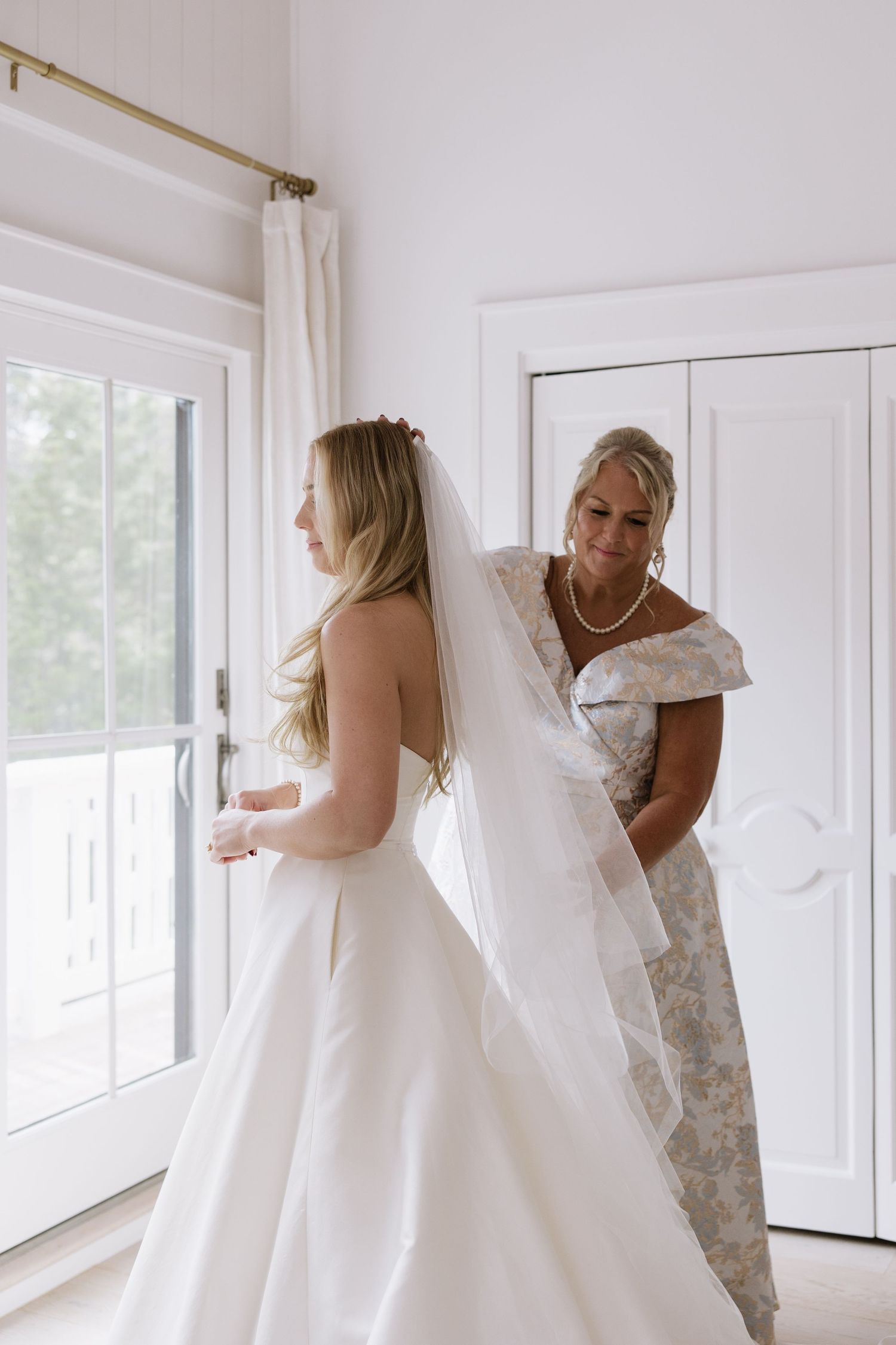 A bride in a white wedding gown gets help adjusting her veil in a bright sunlit room.