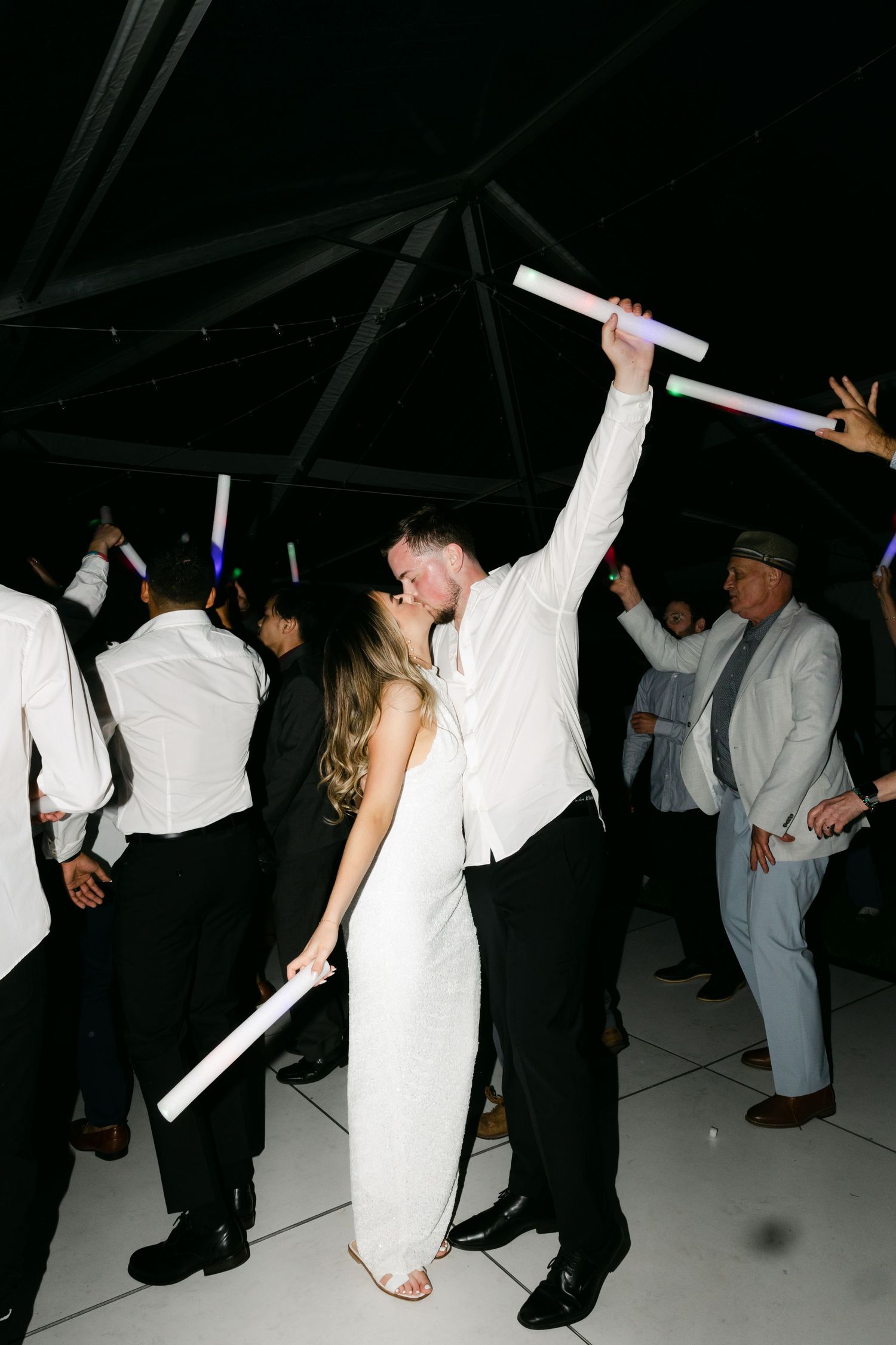 Wedding guests dance and celebrate on a dark dance floor with glowsticks.