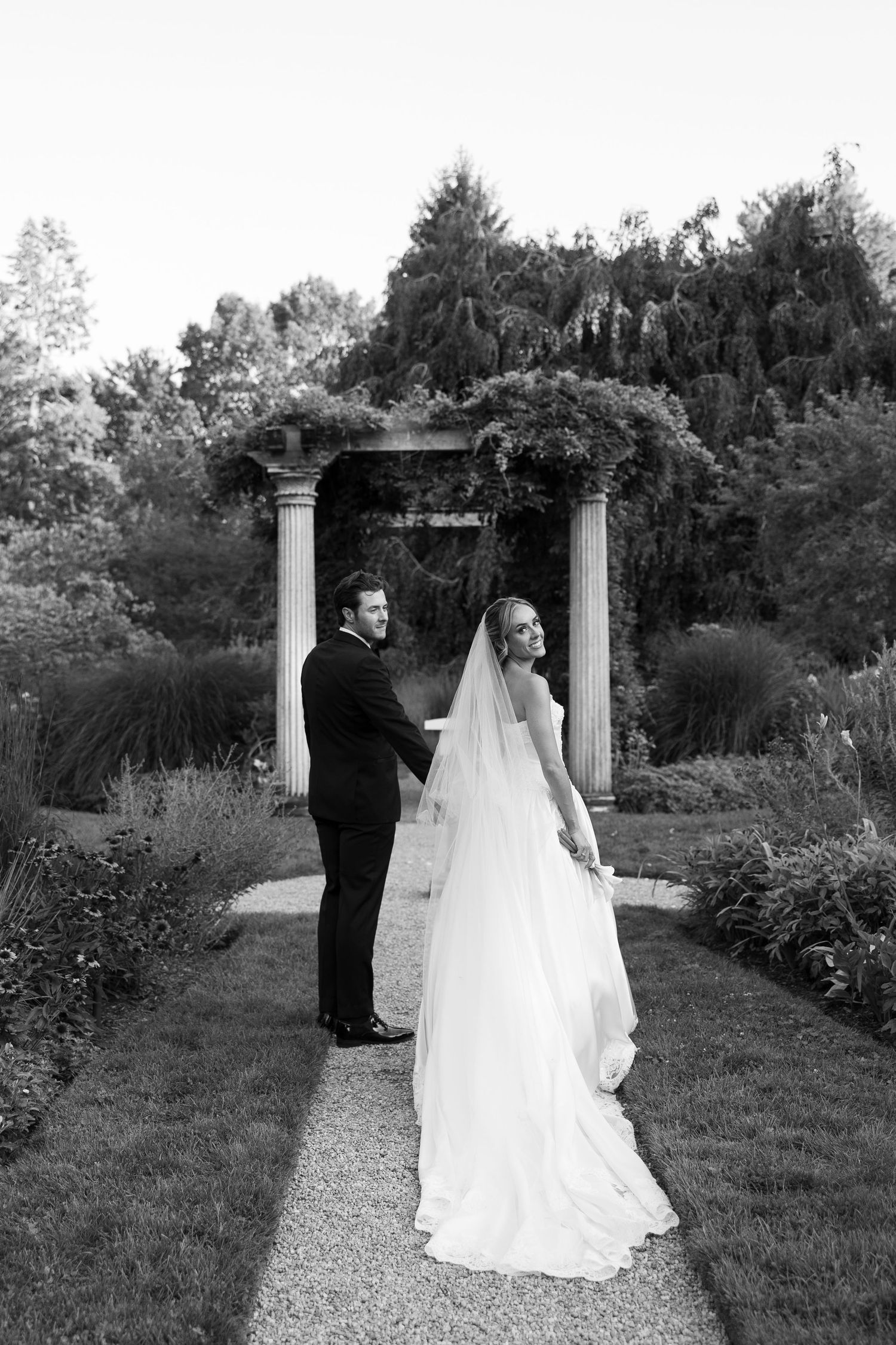 Black and white photo of a couple standing by a garden pergola with a long flowing wedding dress train.