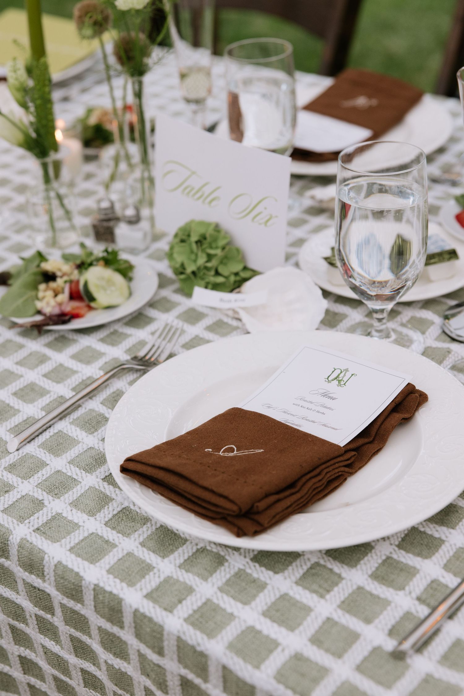 Elegant wedding reception table setting with sage green geometric tablecloth, brown napkin, white plate, and Table Six sign.