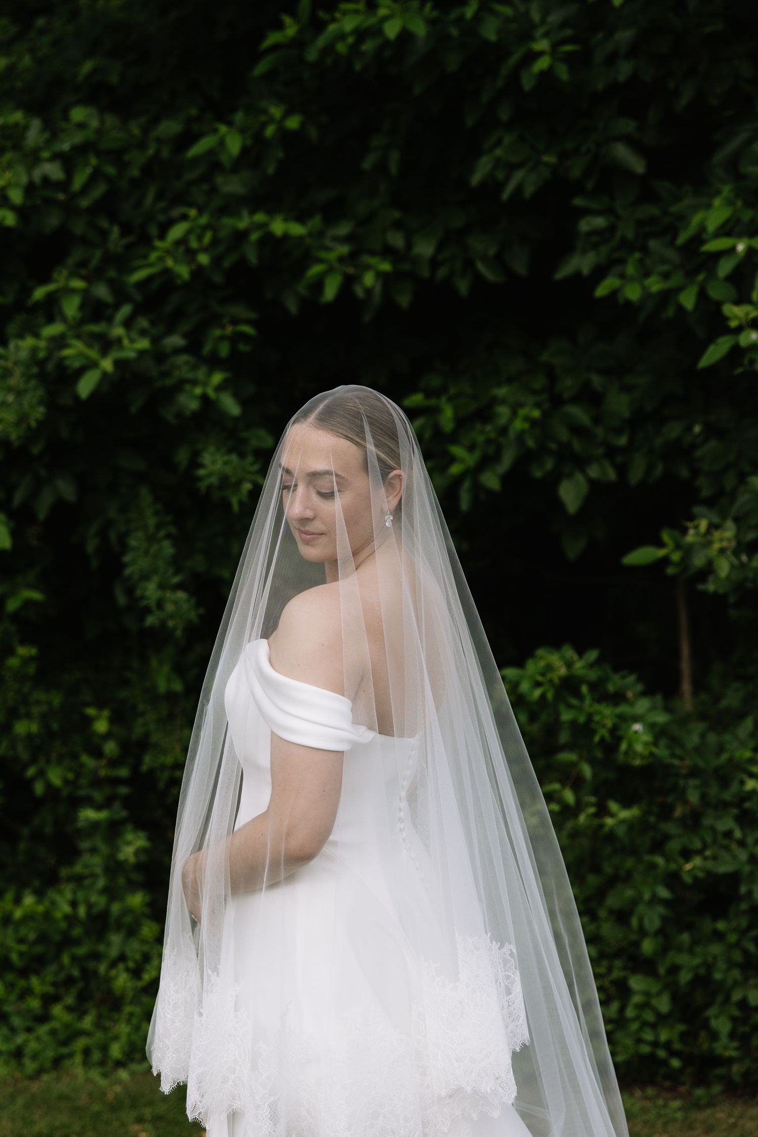 Bride in off-shoulder white gown and long veil poses outdoors against lush green foliage backdrop.