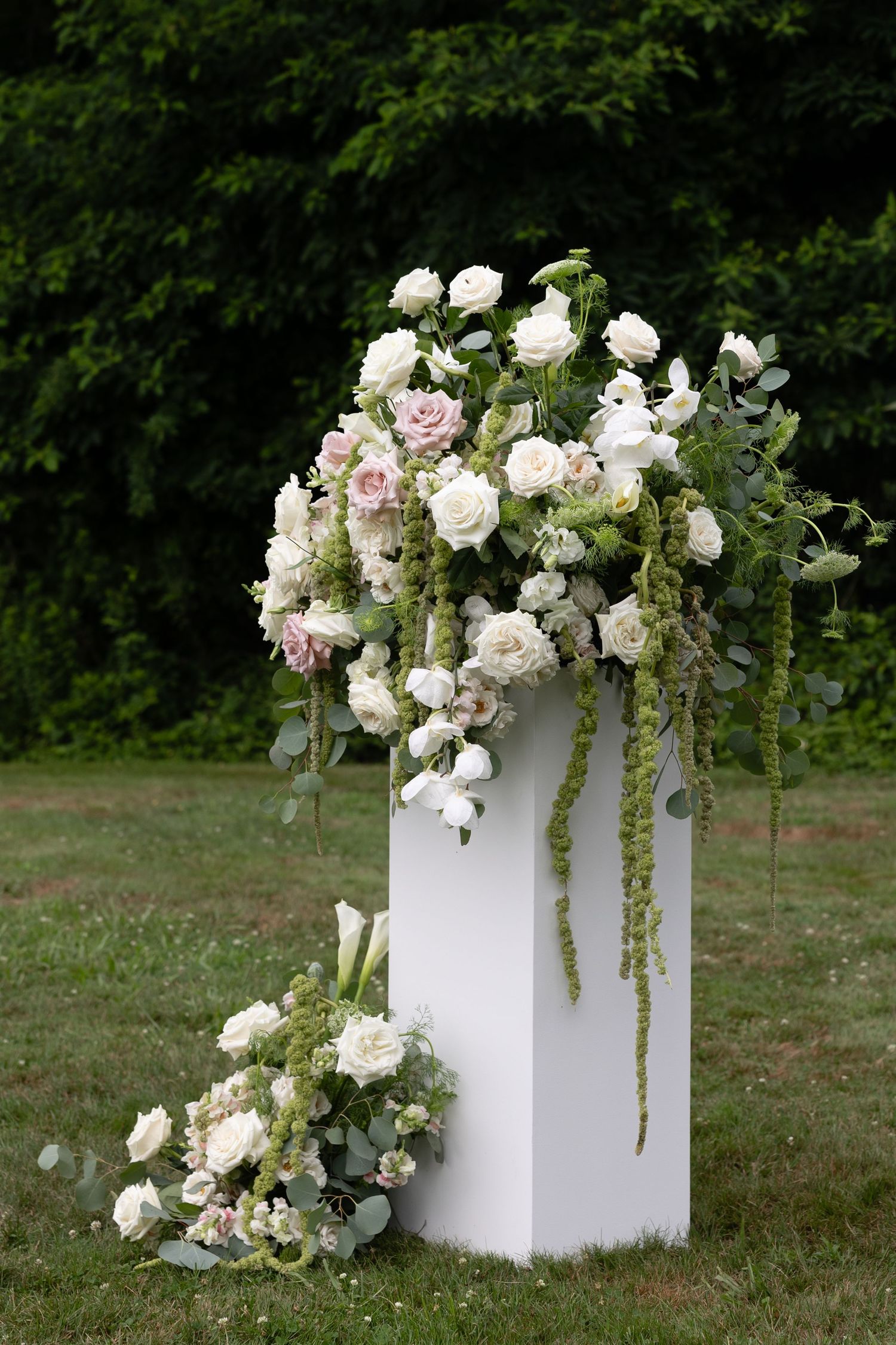 White and blush roses with greenery arranged on a white pedestal outdoors for a wedding ceremony.
