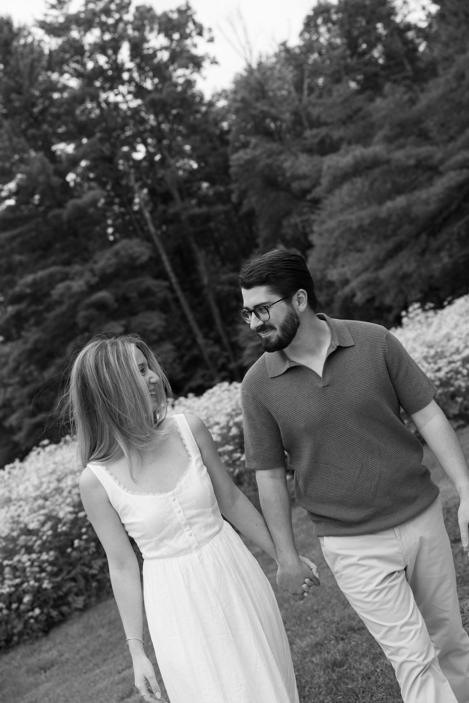Black and white photo of a couple holding hands and smiling at each other while walking outdoors near blooming flowers.