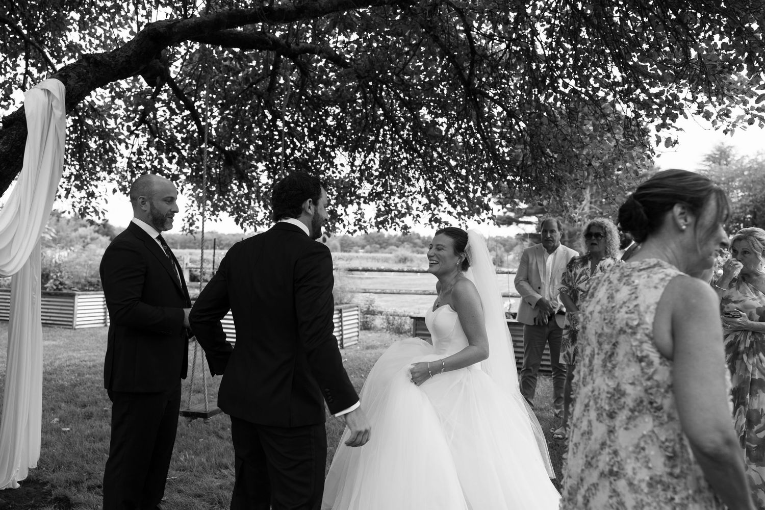 Black and white outdoor wedding ceremony with bride in gown and groom exchanging vows under a tree.