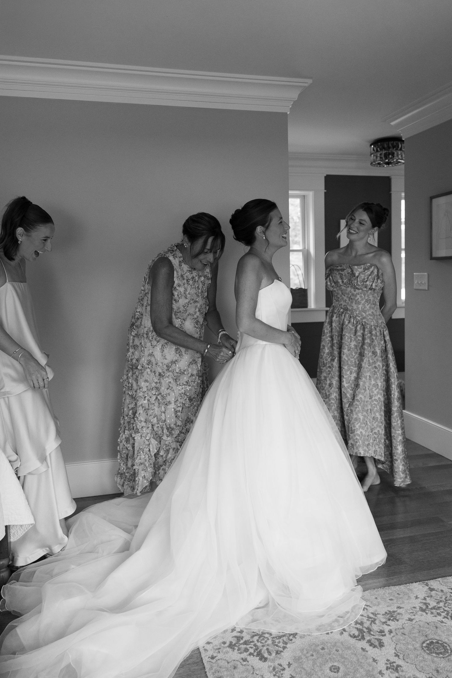 Black and white photo of a bride in a white gown being helped by bridesmaids in floral dresses before the wedding ceremony.