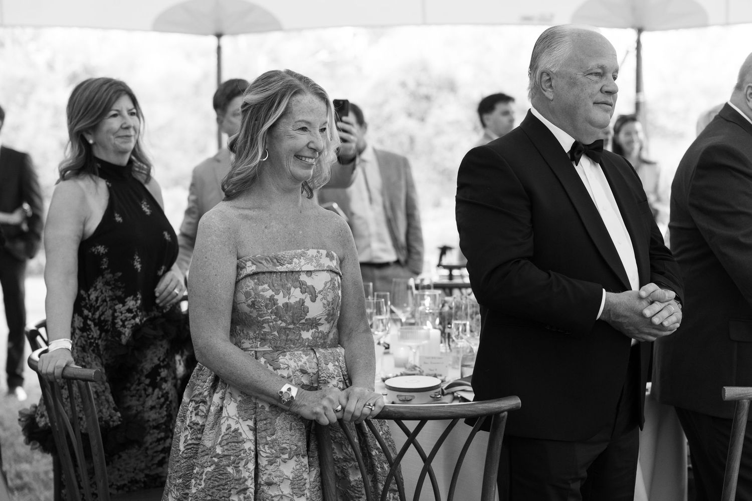 Black and white photo of elegantly dressed guests smiling at a formal outdoor wedding reception under a tent.