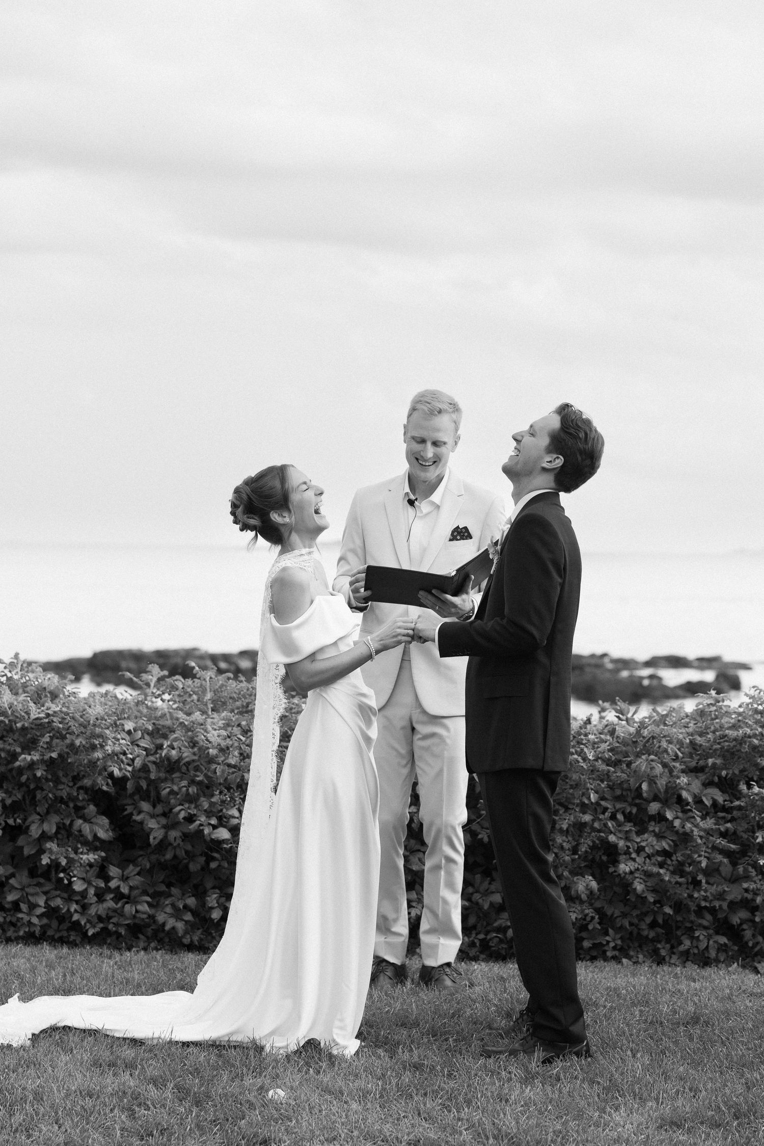 Black and white photo of a bride and groom laughing during outdoor wedding ceremony with officiant.