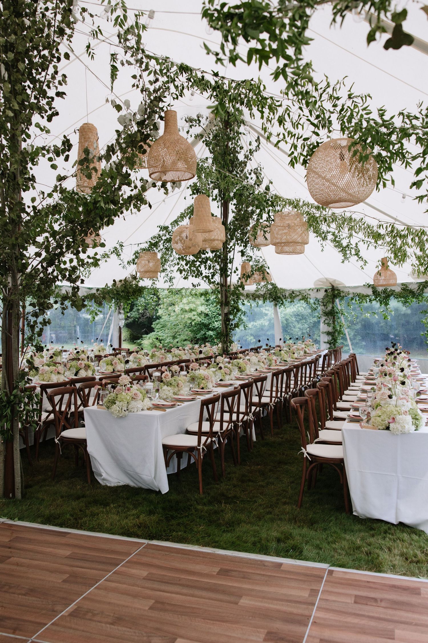 Romantic outdoor wedding reception tent with long banquet tables, wooden chairs, hanging wicker lanterns, and lush greenery decor.