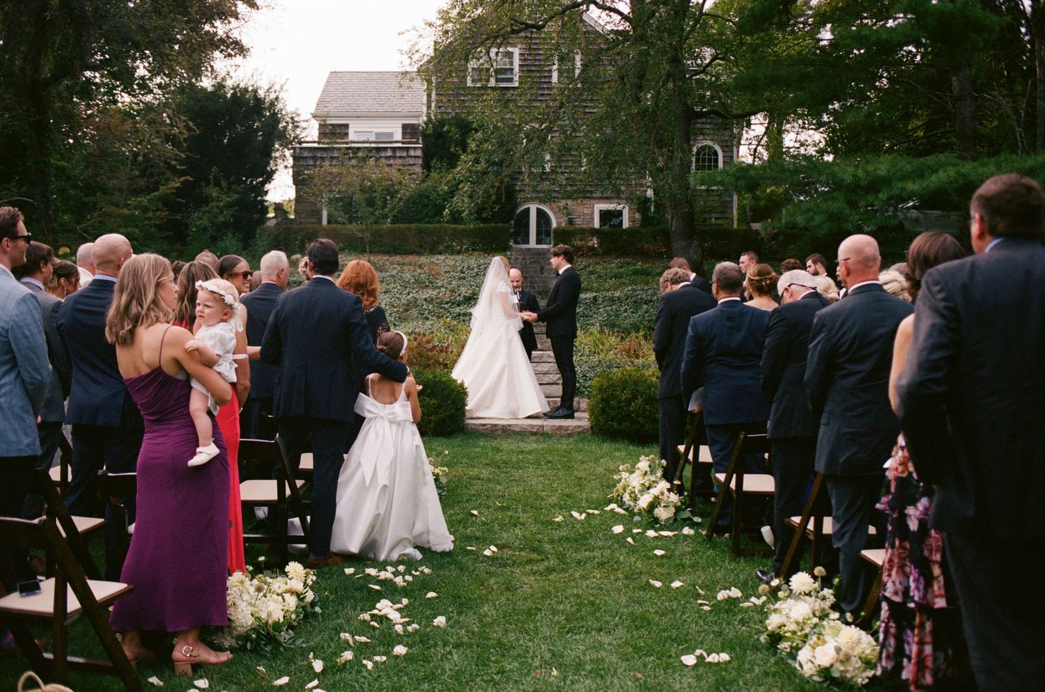 Outdoor wedding ceremony on lush green lawn with guests seated in rows, flower girl, and couple at altar near stone mansion.