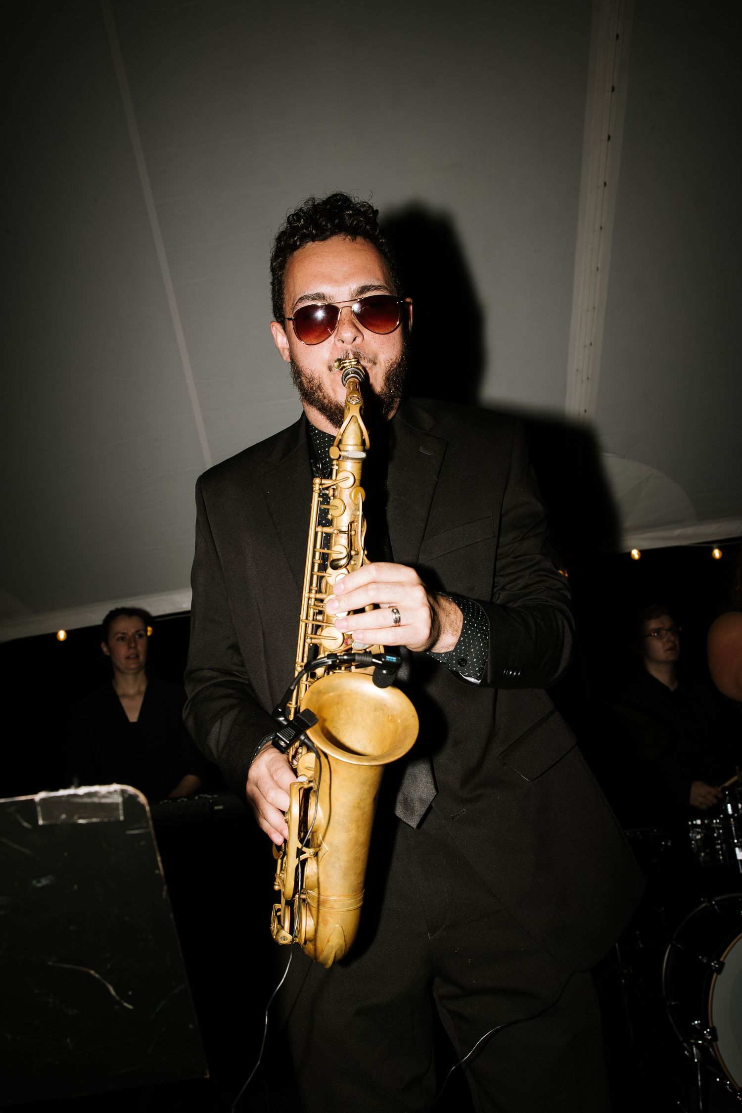 A jazz musician in a black suit and red sunglasses plays alto saxophone at an indoor evening performance.