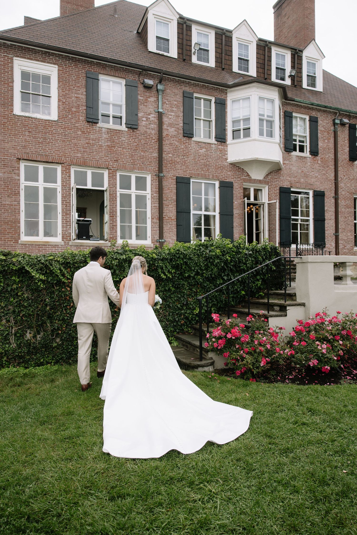 A wedding couple walks along a brick mansion's landscaped grounds with blooming rose bushes.