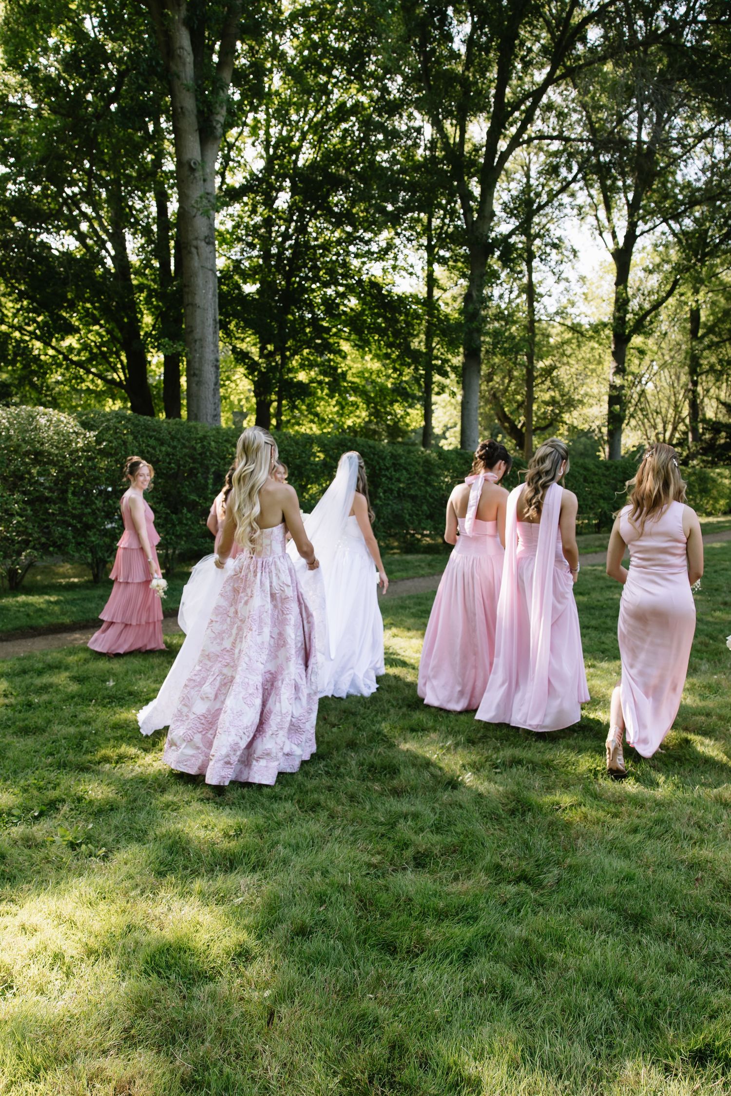 Wedding party in flowing pink dresses walking across sunlit garden lawn.