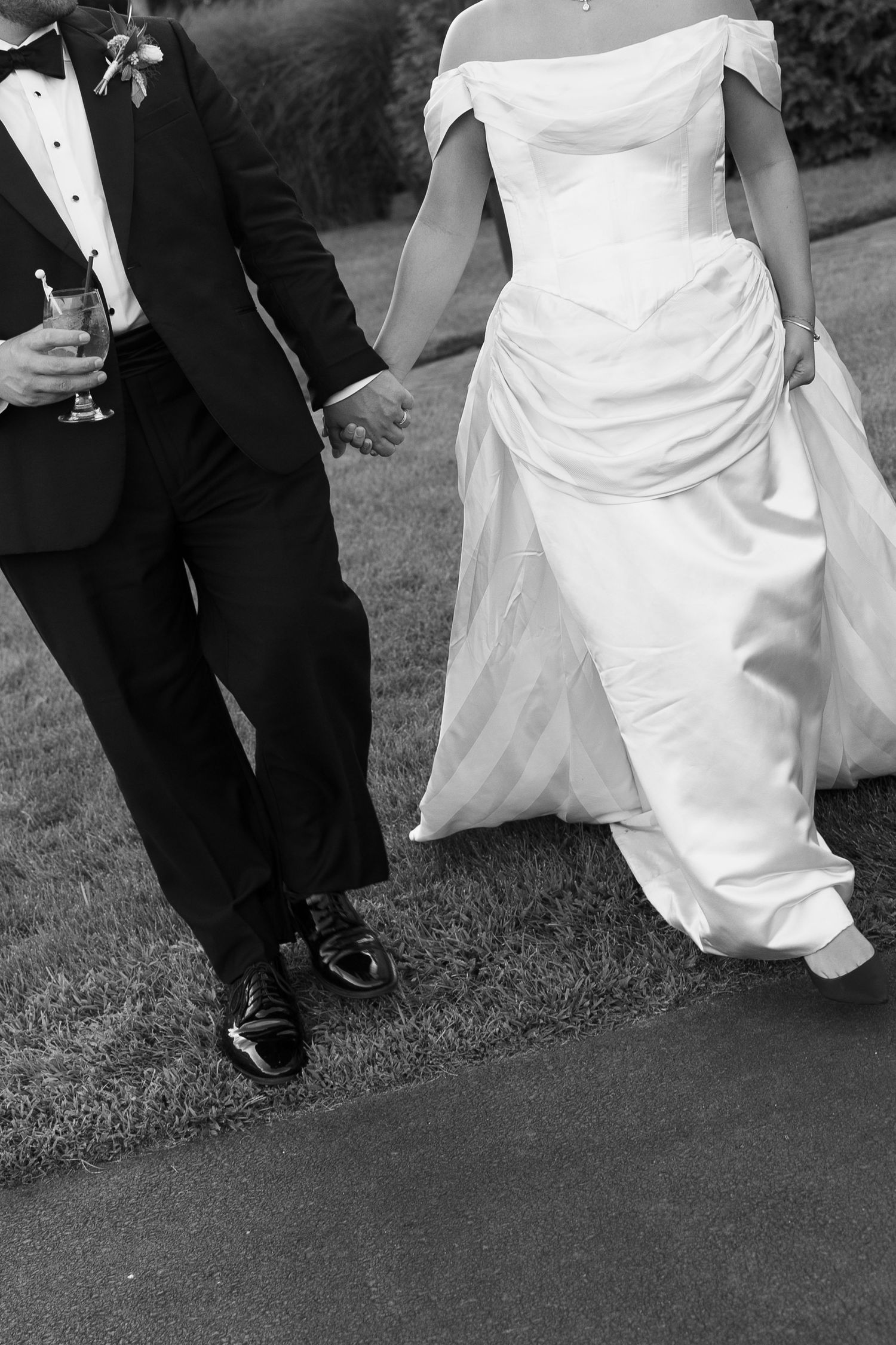 Black and white photo of bride and groom holding hands, walking in elegant wedding attire on a garden path.