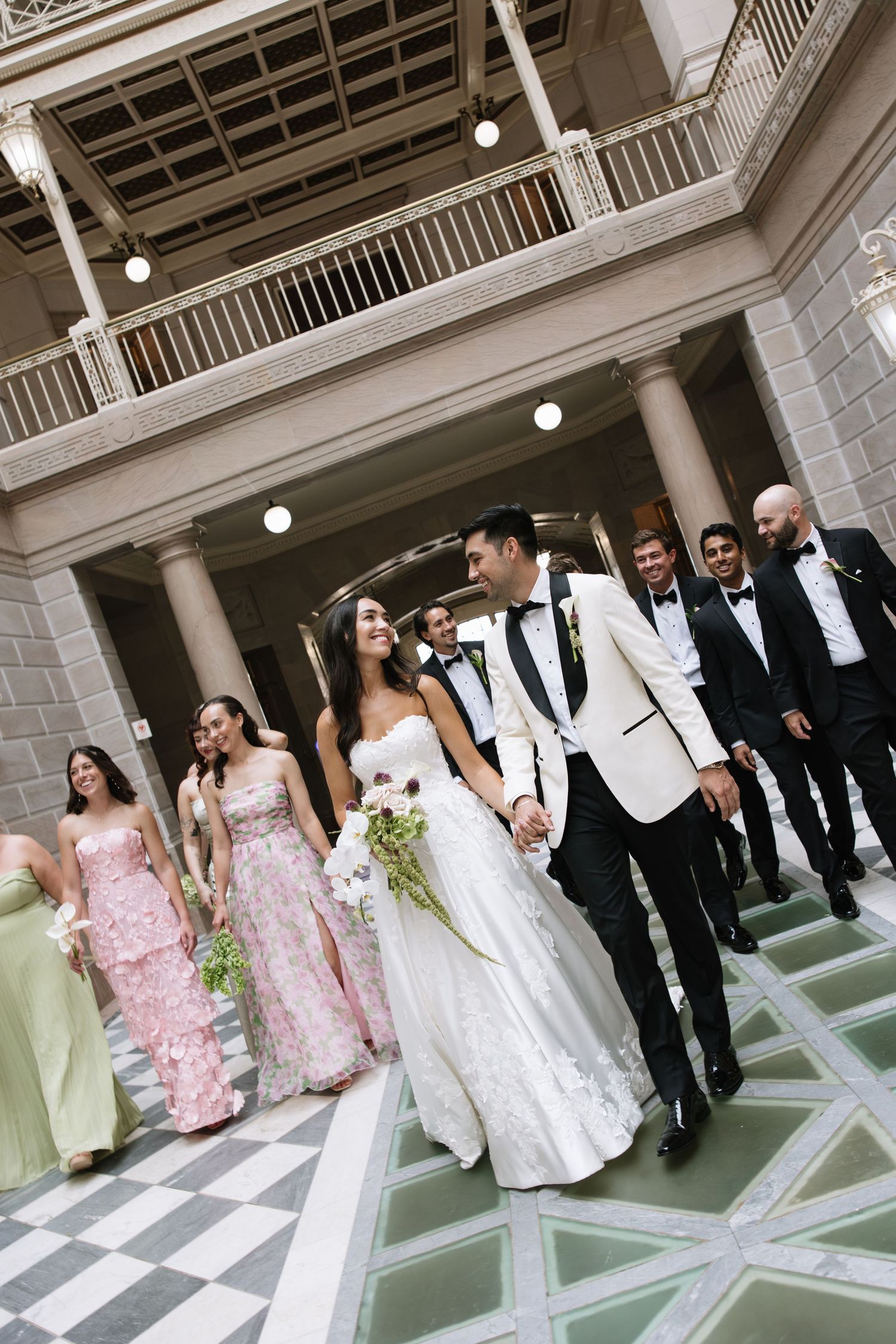 Wedding party walks through elegant columned venue, bride in white gown holding bouquet with bridesmaids in pink and green dresses.