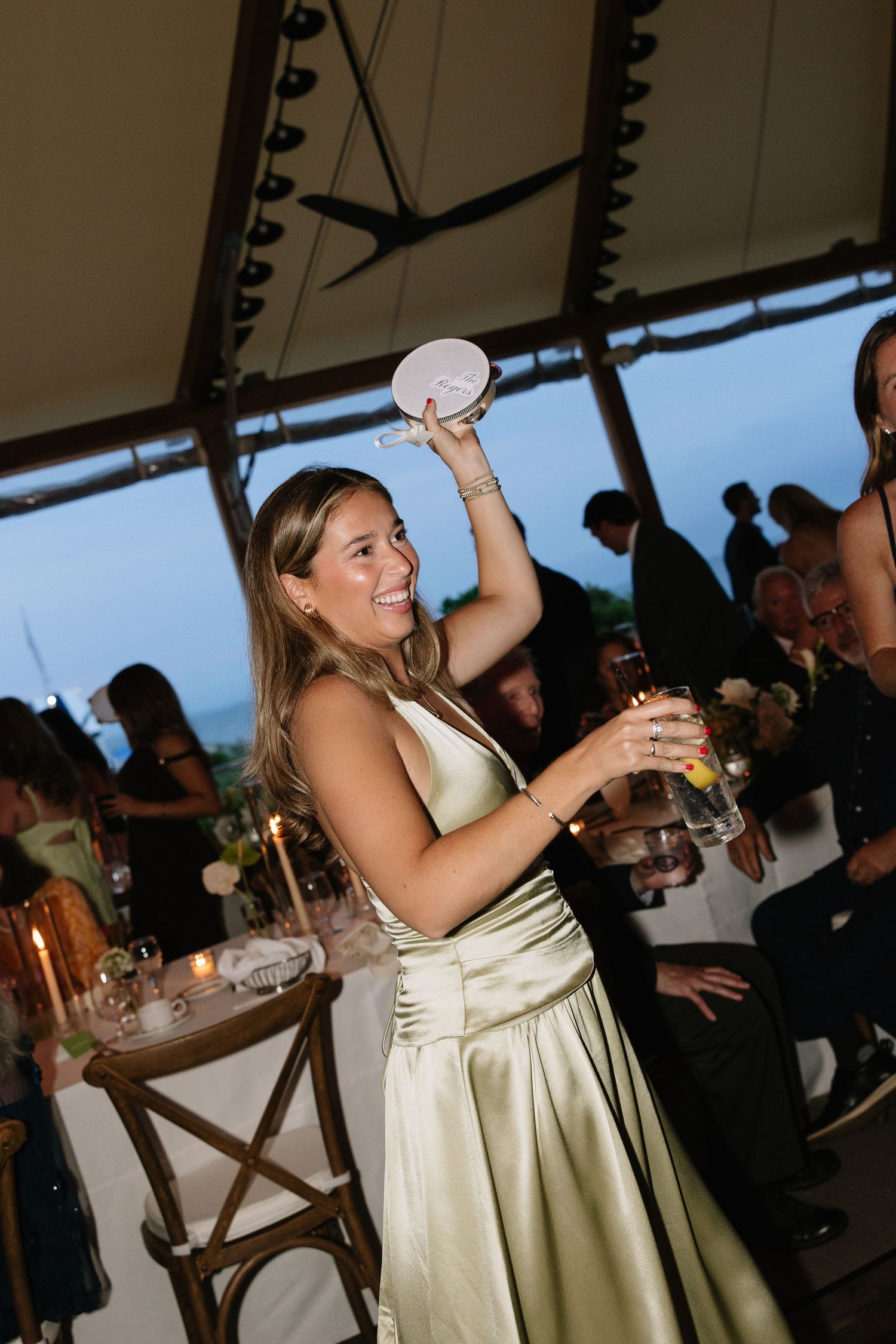 A smiling woman in a sage green satin gown raises a glass at a festive outdoor wedding reception under a tent.