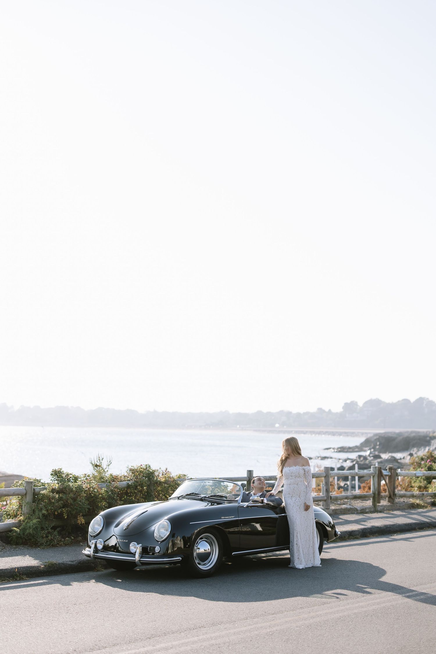 Bride in white gown posing beside a vintage black Porsche 356 convertible on a coastal road with ocean views.