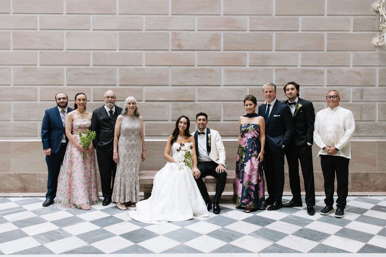 Wedding party poses in elegant venue with checkered floors, bride in white gown seated beside groom in tuxedo.