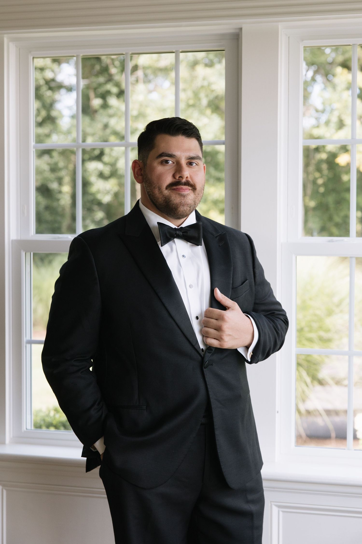 Groom in black tuxedo with bow tie poses confidently near white-framed windows with garden views.