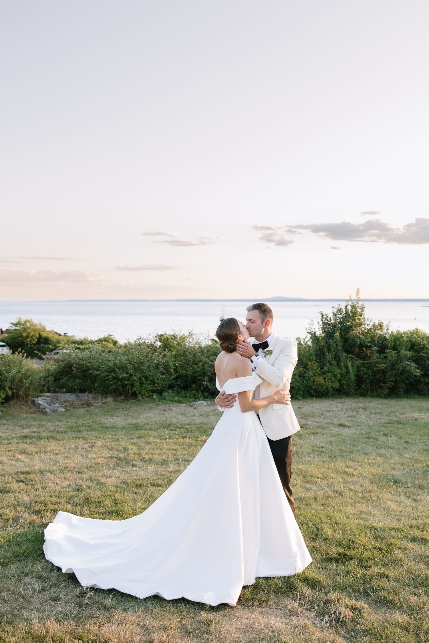Bride and groom embracing at sunset by the ocean, bride in off-shoulder gown with long train.