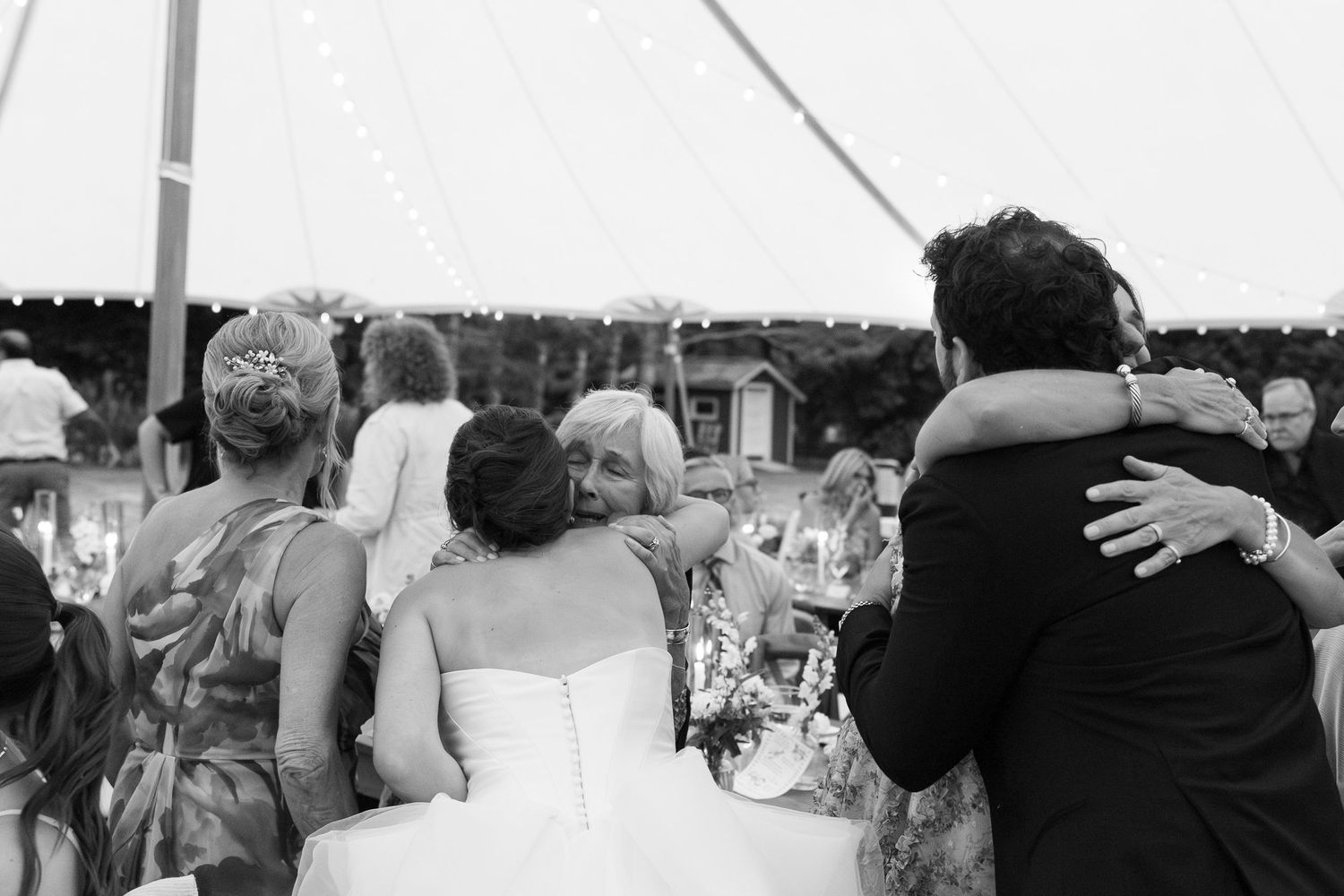 Black and white photo of bride and groom sharing warm hugs with elderly wedding guest under tent.