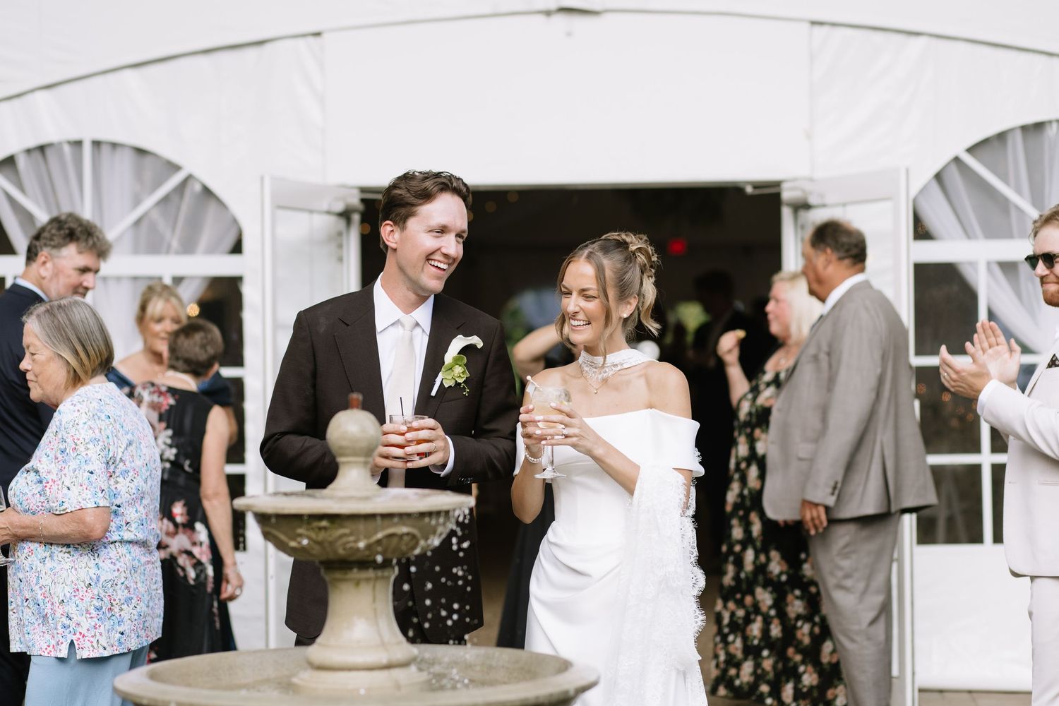 Newlywed couple laughing together outside a white wedding tent, bride in off-shoulder gown.