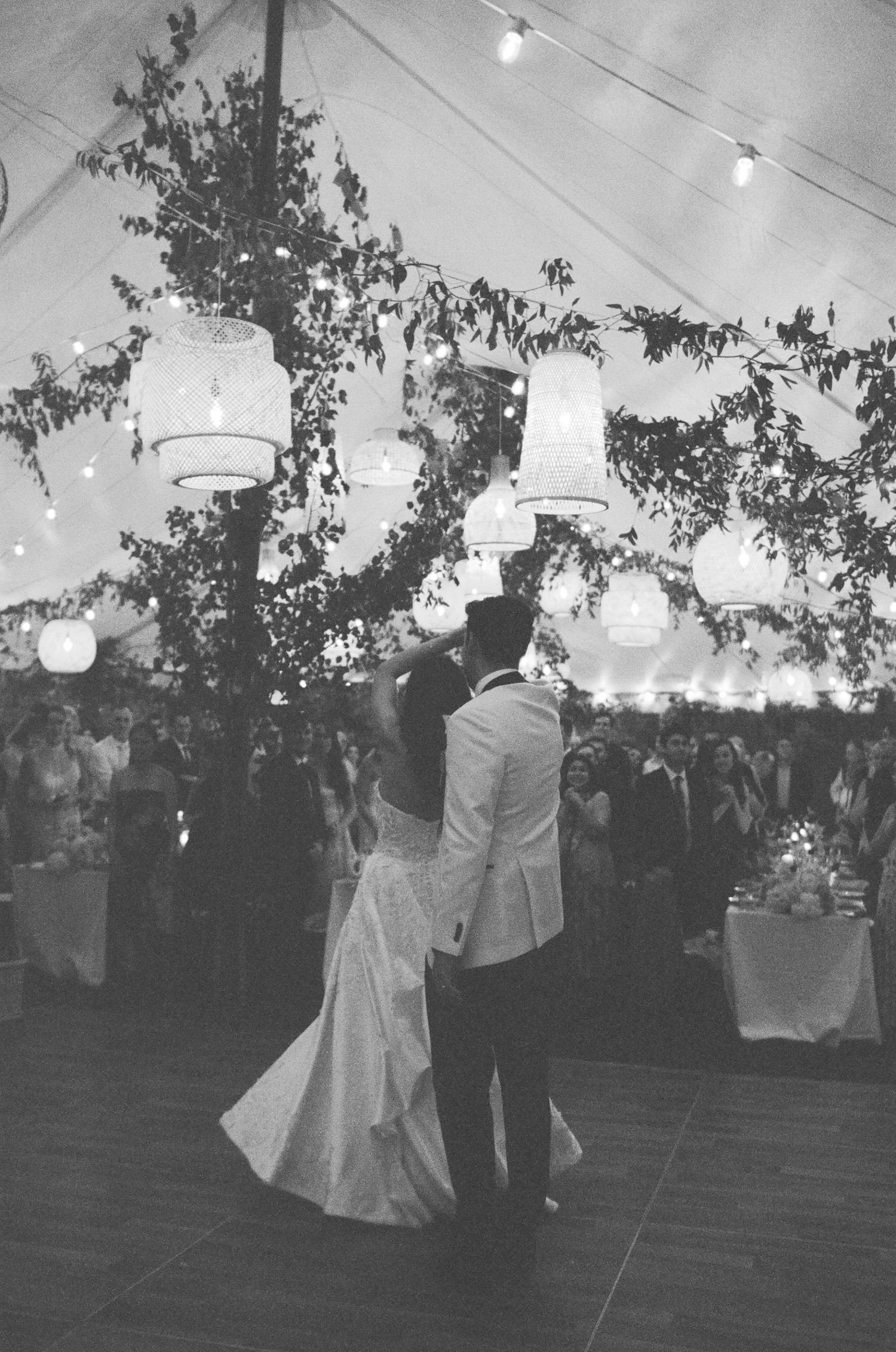 Black and white photo of bride and groom sharing first dance under glowing lanterns and string lights at elegant wedding reception.