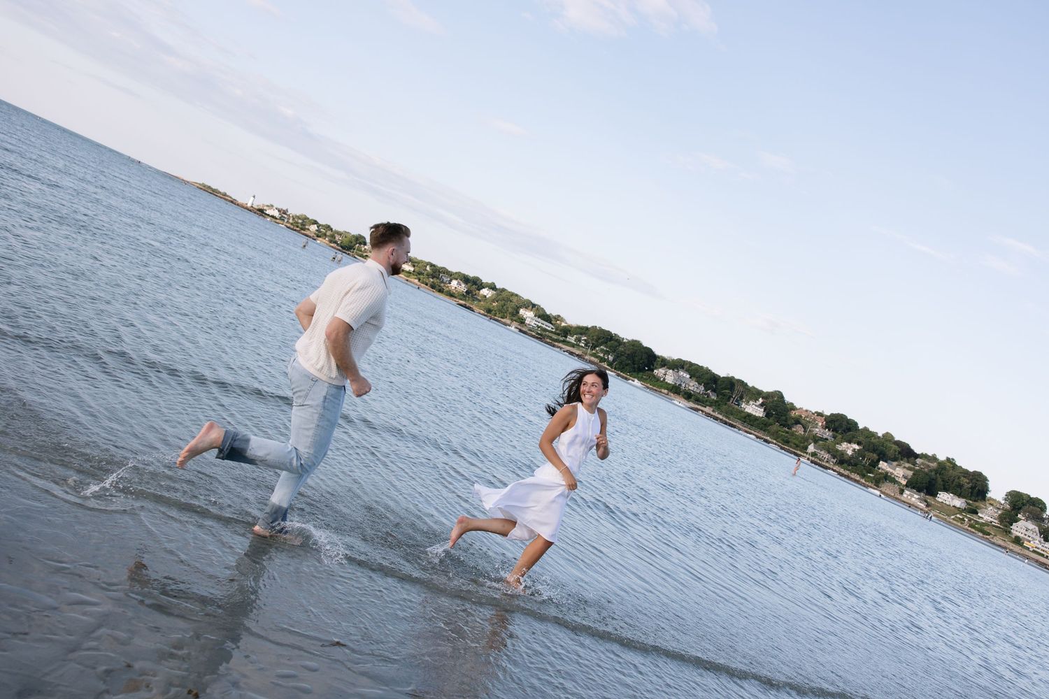 Couple running playfully through shallow ocean water on a sunny beach, wearing white casual clothes.