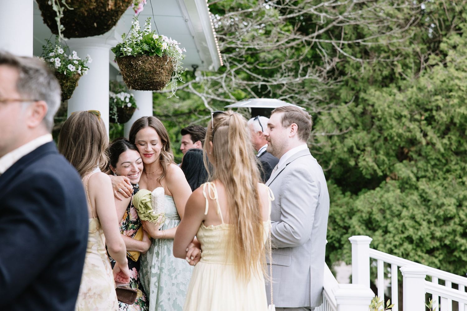 Wedding guests celebrate on a white porch, bridesmaids in yellow dresses laughing with the groom in a gray suit outdoors.