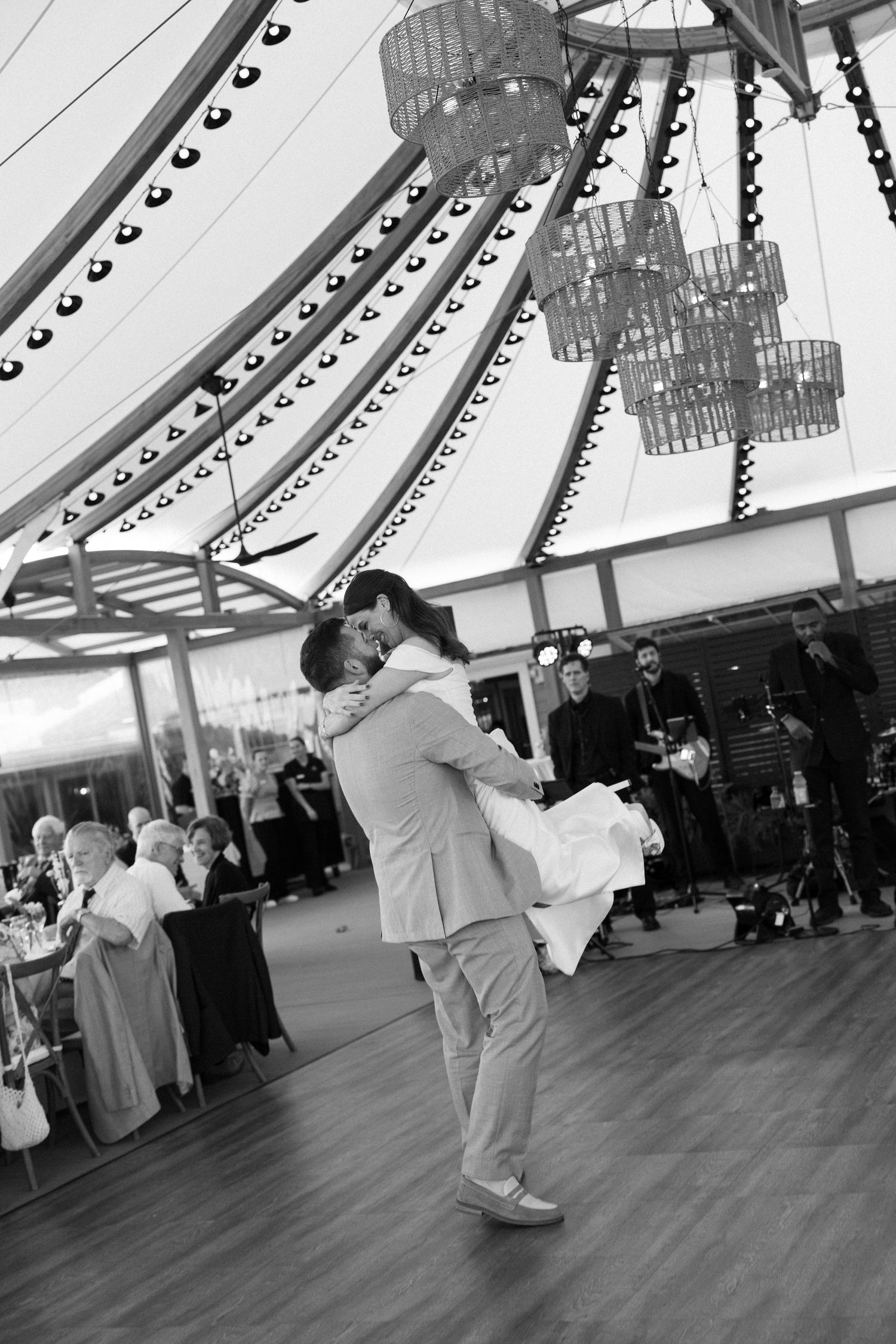 Groom lifts bride during first dance at elegant tented wedding reception in black and white photo.