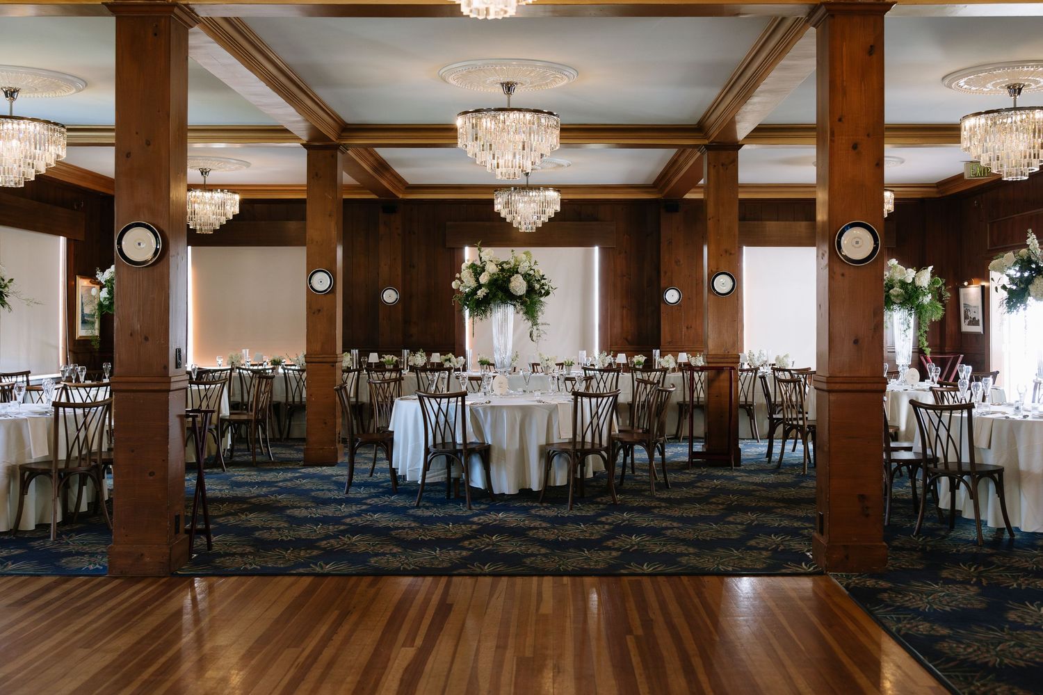 Elegant banquet hall with wood columns, crystal chandeliers, round tables with white linens, and floral centerpieces on patterned carpet.