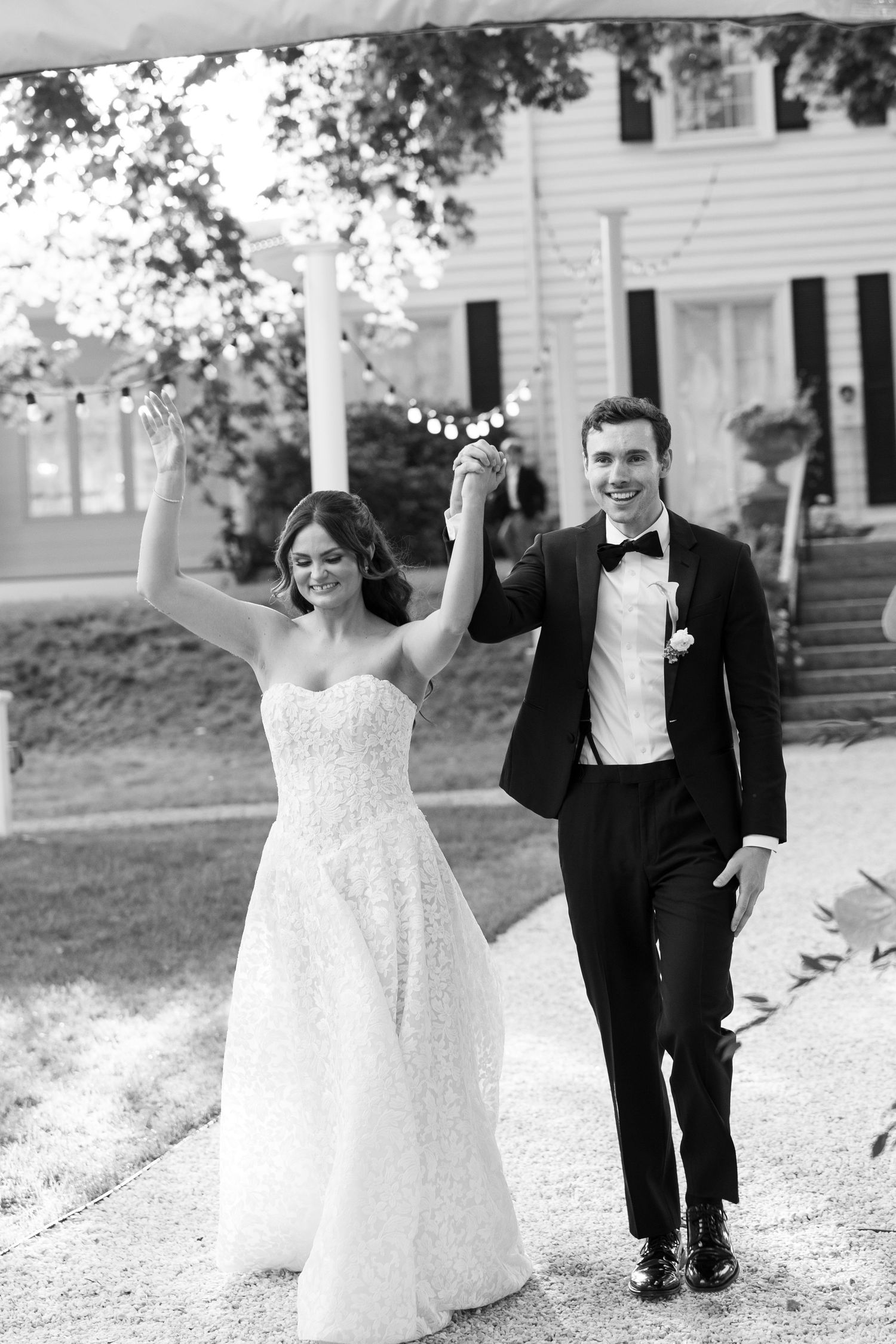 Black and white photo of a joyful bride and groom holding hands raised, walking together outside a white colonial home.