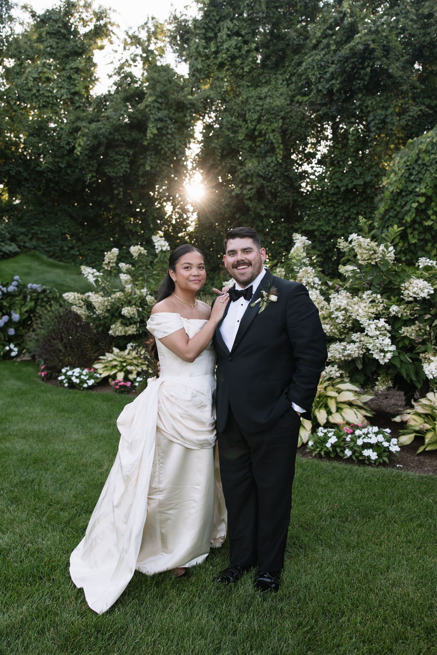 Smiling bride and groom pose together in lush garden at golden hour.