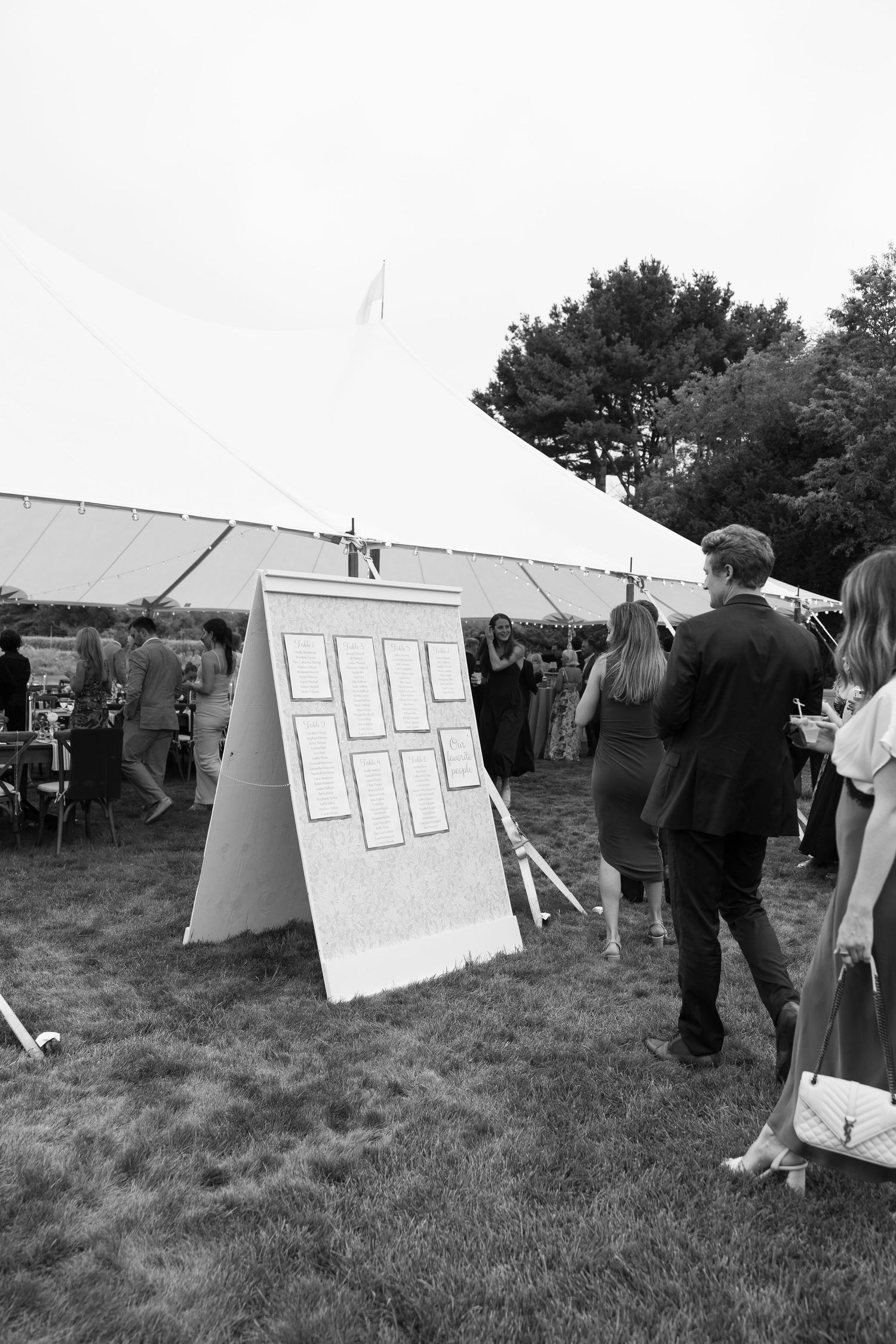Wedding guests browse seating chart display board outside large white tent at outdoor reception.