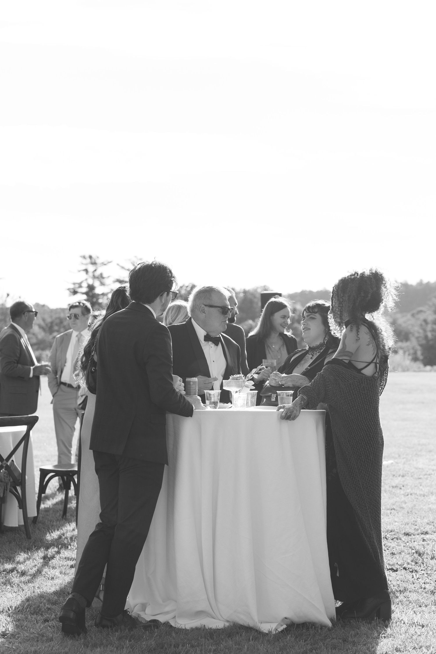 Wedding guests mingle around cocktail table at outdoor reception in elegant black and white photo.