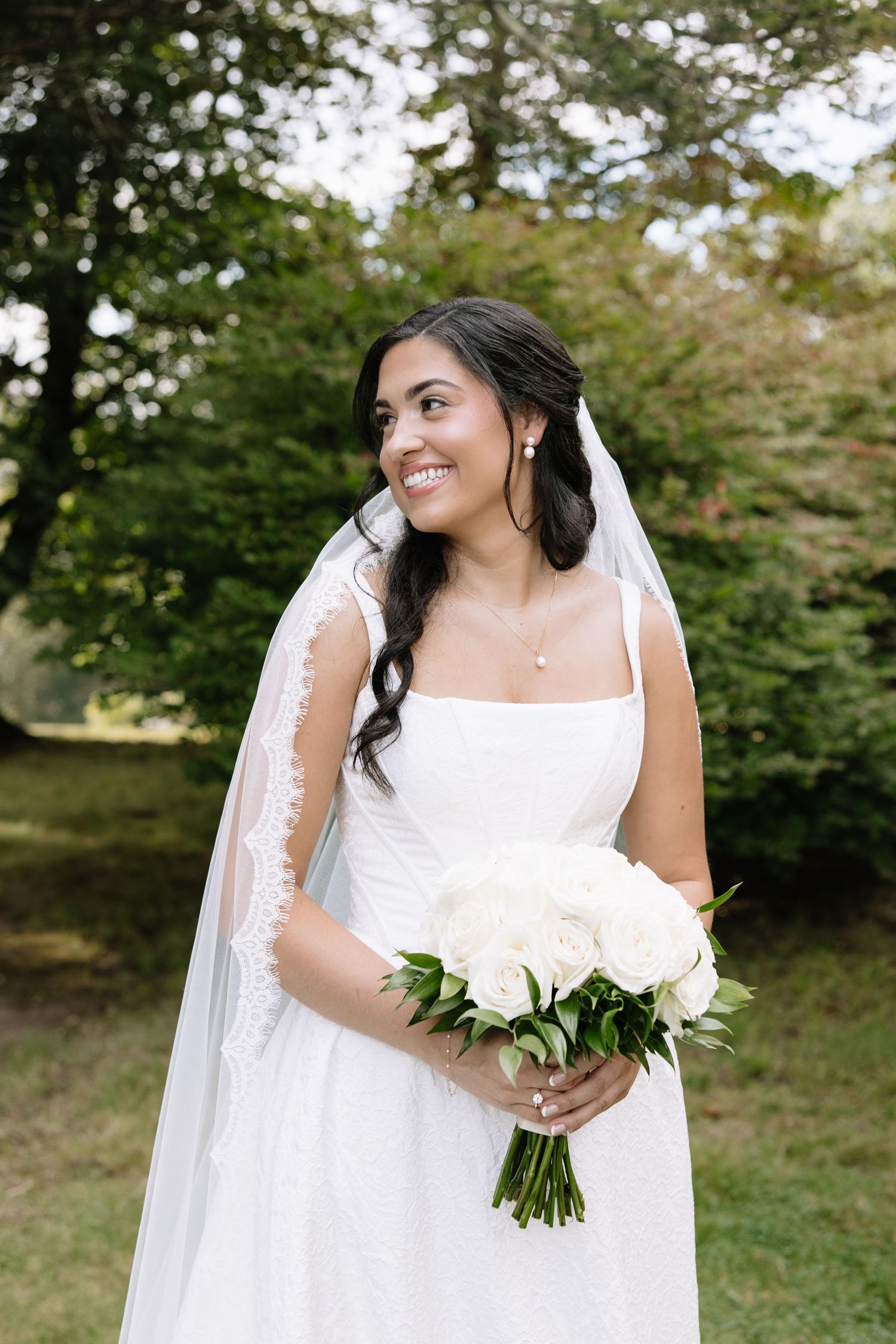 Smiling bride in white gown and veil holds white rose bouquet outdoors among lush green trees.