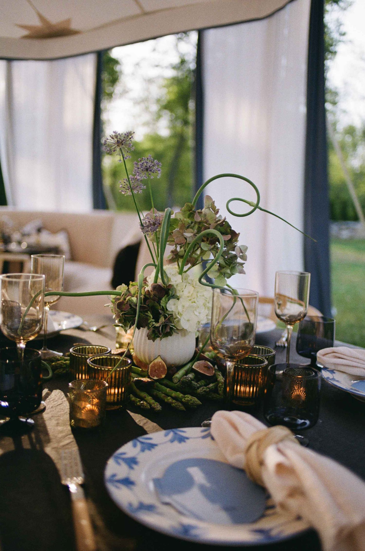 Elegant wedding table setting with floral centerpiece, asparagus, blue and white plates, and amber votives under a tent.