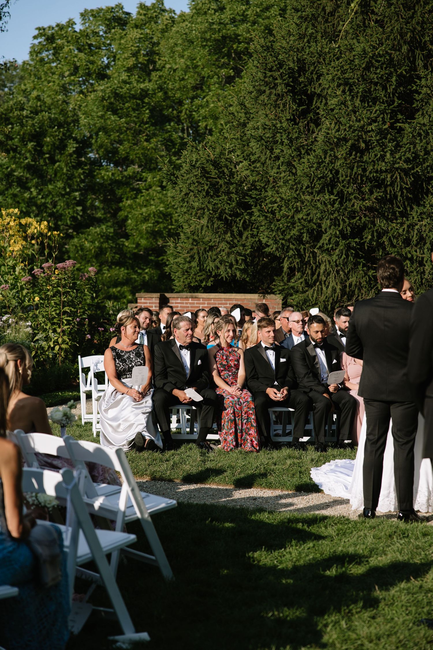 Wedding guests seated on white chairs watch an outdoor garden ceremony at sunset.